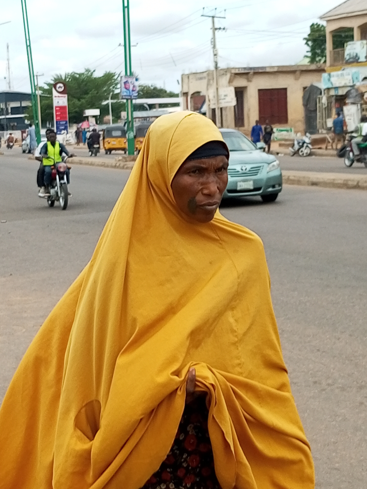 A woman wearing a yellow hijab walks on a paved street in Tempure, Nigeria. She has dark skin and is wearing a black scarf under her hijab. A small patch of green makeup is visible on her cheek. She is draped in a large yellow fabric, and a floral pattern is visible on her clothing beneath it. In the background, a blue sedan drives by, and further in the distance, a yellow auto-rickshaw is visible. A man wearing a neon green vest rides a motorcycle in the left background. Buildings with tan and white facades line the street. Power lines and a traffic light are visible overhead. The sky is overcast.