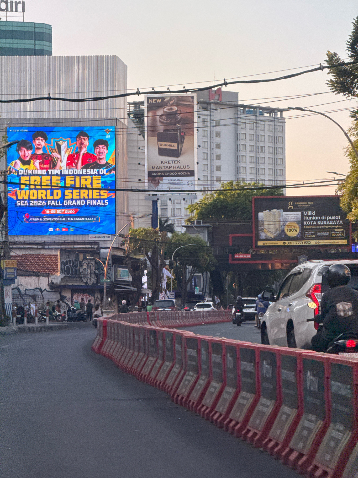 The image depicts a street scene in Surabaya, Indonesia, characterized by a busy road and several billboards. In the foreground, a street with a dark asphalt surface is bordered by a series of red and white plastic barriers. Several vehicles, including cars and motorcycles, are visible on the road, with some appearing to be stationary. A few individuals are seen on motorcycles, some wearing helmets.
Dominating the background are multiple large billboards. One prominent blue billboard displays an advertisement for "FREE FIRE WORLD SERIES SEA 2026 FALL GRAND FINALS," featuring images of four young men. To the right of this billboard, another billboard advertises "DUNHILL KRETEK MANTAP HALUS AROMA CHOCO MOCHA." Further to the right, a black billboard promotes "HUNIAN di PUSAT KOTA SURABAYA," an apartment complex with a depicted building and text indicating "FREE DP 100%."
The environment includes multi-story buildings, some with modern architecture and others appearing older, with visible windows and signage. Trees with green foliage are also present, and power lines are strung across the scene. The sky is overcast, suggesting it is either early morning or late afternoon. The overall impression is that of an urban commercial area.