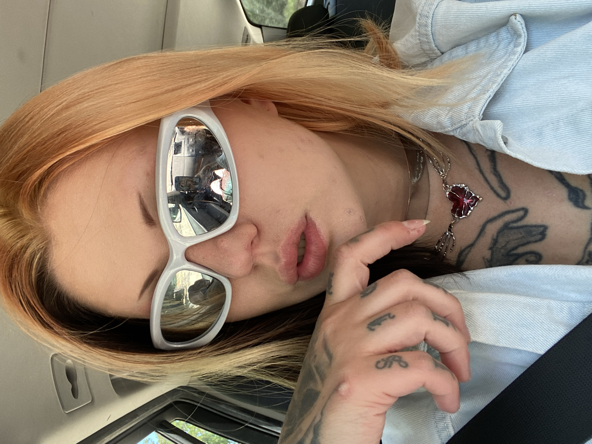 A woman is sitting in a car, facing towards the camera and tilting her head to the left. She is wearing white-rimmed sunglasses, which reflect the inside of the car and exterior scenery. She has light orange hair and is wearing a light blue denim jacket. A black seatbelt is visible across her body. The woman has tattoos on her hand, neck, and chest. She is wearing a silver necklace with a red floral pendant. The location is inside a car, potentially driving through or parked in Kruče - Kërruçi, Montenegro, as suggested by the prompt. The reflection in the sunglasses likely shows the surrounding environment.