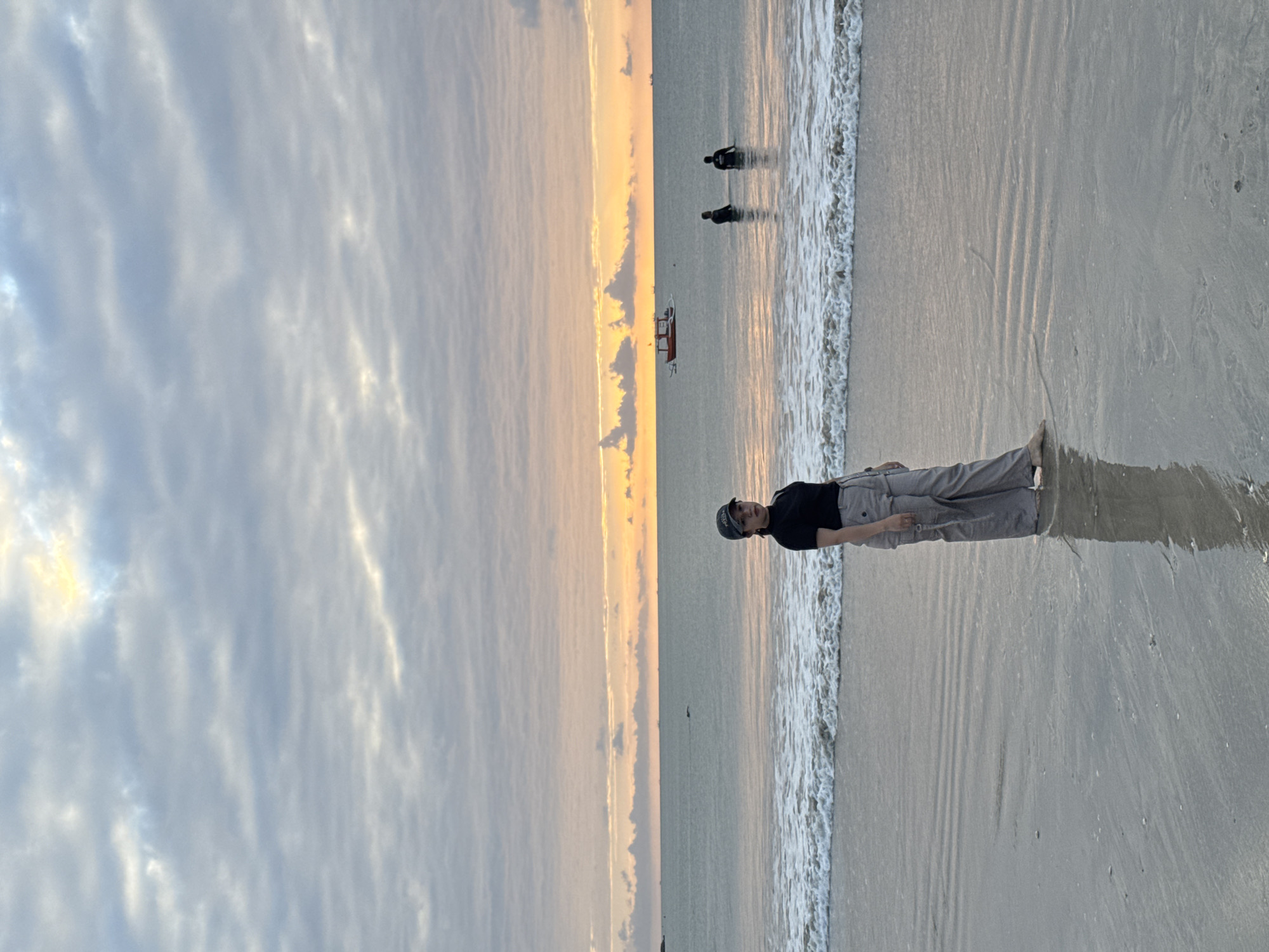A woman stands on a beach in Kuta Selatan, Indonesia, facing the ocean. She is wearing a black t-shirt, grey pants, and a dark baseball cap. The tide is coming in, with gentle waves lapping at the shore. In the background, the ocean stretches to the horizon, and a few distant boats are visible. The sky is filled with soft, grey clouds, with streaks of orange and yellow indicating either sunrise or sunset. The overall mood of the image is peaceful and serene.