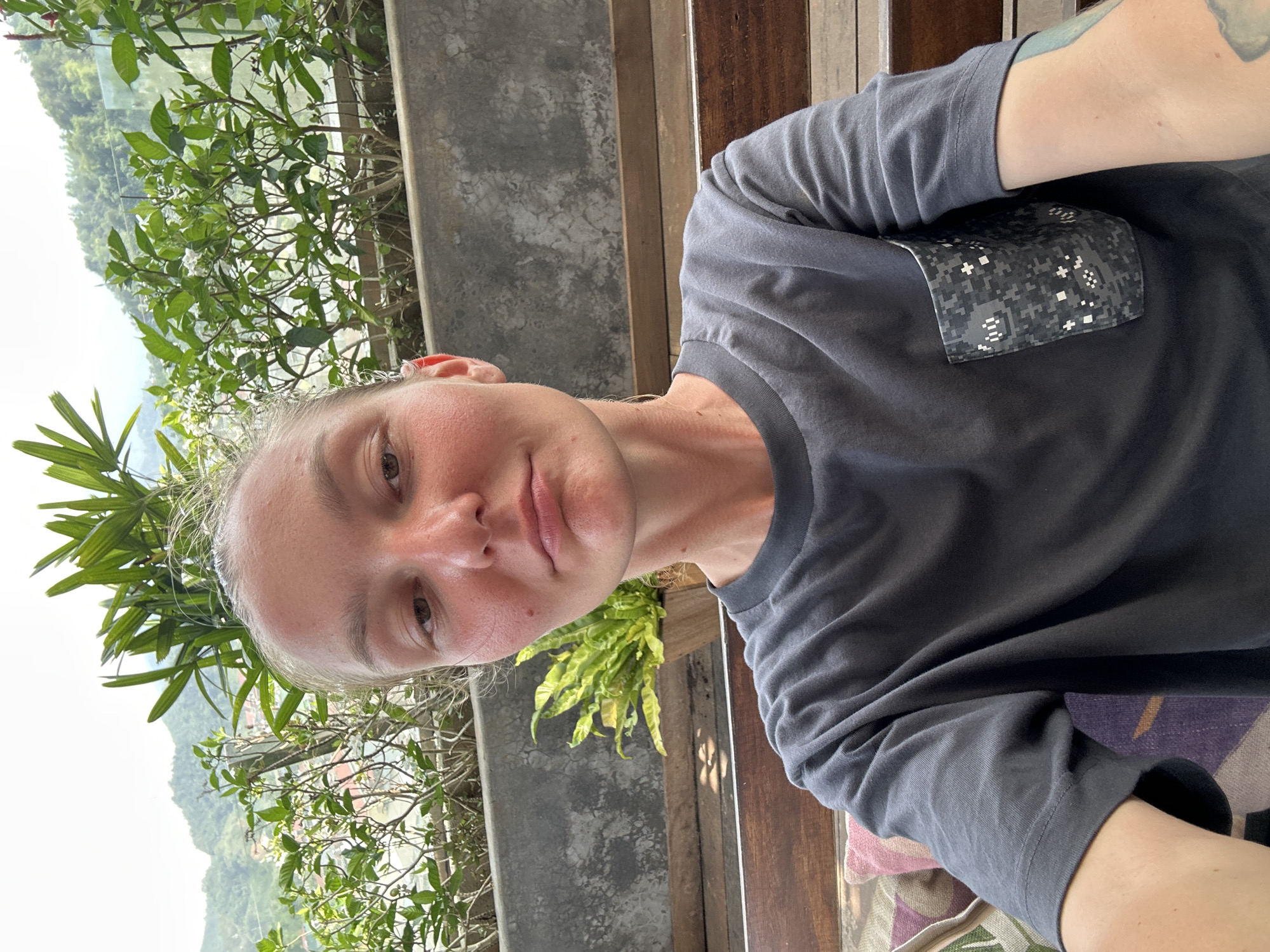 The media file displays a close-up selfie of a person outdoors, with the image oriented on its side. The subject, appearing to be female with light skin and light-colored hair pulled back, looks directly at the camera with a neutral expression. She wears a dark gray short-sleeved t-shirt featuring a unique pixelated digital camouflage pocket on the left chest. A tattoo is visible on her left arm.
The setting behind the person is a verdant, mountainous landscape under a bright sky, characteristic of a daytime view in a location like Hà Giang 1 Ward, Vietnam. Dense green foliage, including bushes with small white flowers and broad palm-like leaves, is prominently featured in the mid-ground. Elements of concrete and wooden structures, possibly a balcony railing or furniture, frame the scene. A glimpse of colorful, patterned fabric, perhaps a cushion or blanket, is visible in the lower right portion of the frame. The overall impression is one of natural beauty and an outdoor, possibly elevated, vantage point.