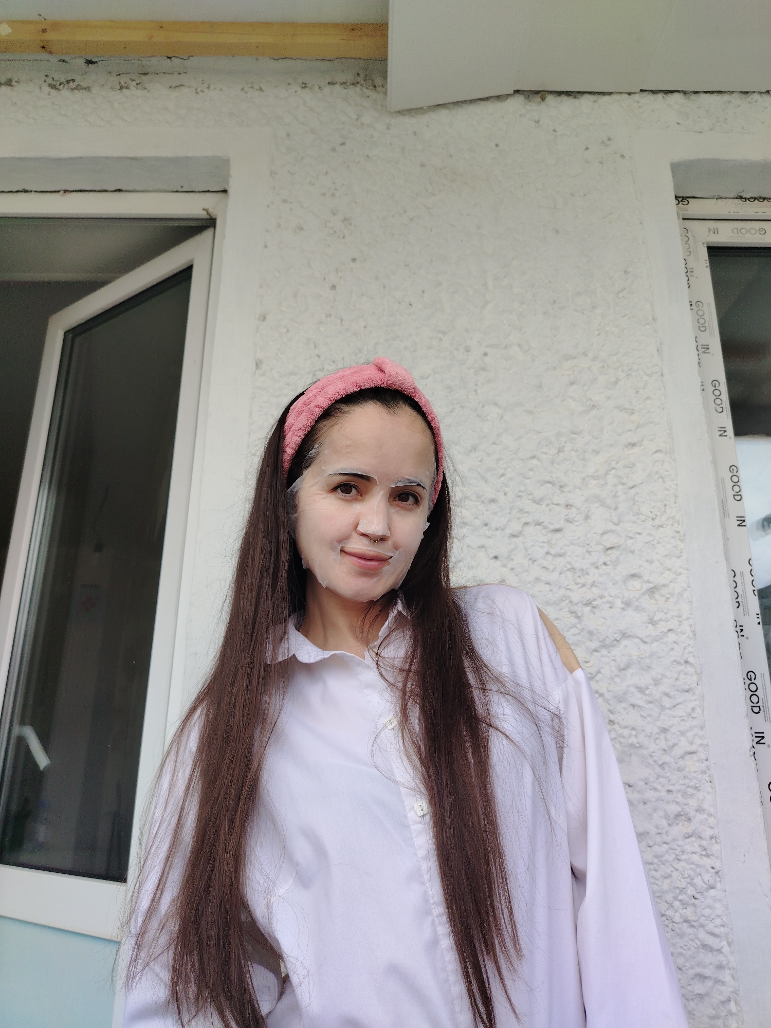 A young woman stands facing the viewer, captured in a brightly lit, medium-shot portrait. She has long, dark brown hair that flows over her shoulders and down her chest. On her head, she wears a pink, knotted headband that keeps her hair off her forehead. Her face is covered with a white sheet mask, which has cutouts for her eyes, nose, and mouth. She looks directly at the camera with a calm expression and a subtle, pleasant smile.
She is wearing a white, long-sleeved collared shirt. Behind her is a textured, white wall. To her left, part of an open white-framed window is visible. To her right, another white-framed window is present, and its vertical frame distinctly features the words "GOOD IN" repeated several times in dark lettering. Above the windows, a rough wooden beam runs horizontally across the top of the frame, indicating structural elements. The lighting suggests it is daytime. The setting appears to be an indoor space or a covered outdoor area like a balcony, likely in Noviy, Russia.