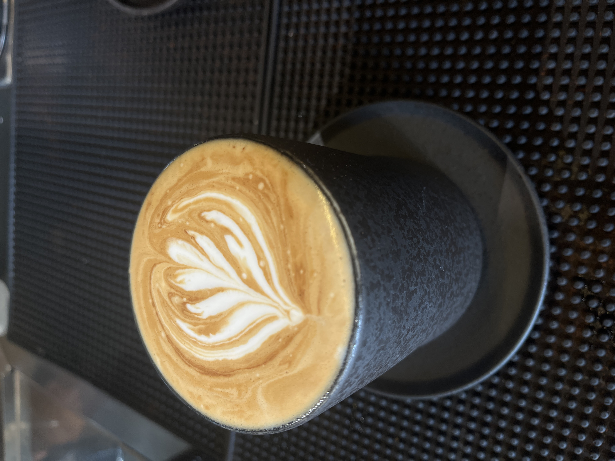 An overhead shot shows a latte with intricate latte art, in a dark grey ceramic cup, on a textured black surface. The latte art forms a delicate pattern of white foam against the rich brown of the coffee. The cup is resting on a matching saucer. The background is blurred, but appears to be a countertop in a cafe, possibly in North Kuta, Indonesia. The lighting is warm and inviting, highlighting the creamy texture of the coffee.