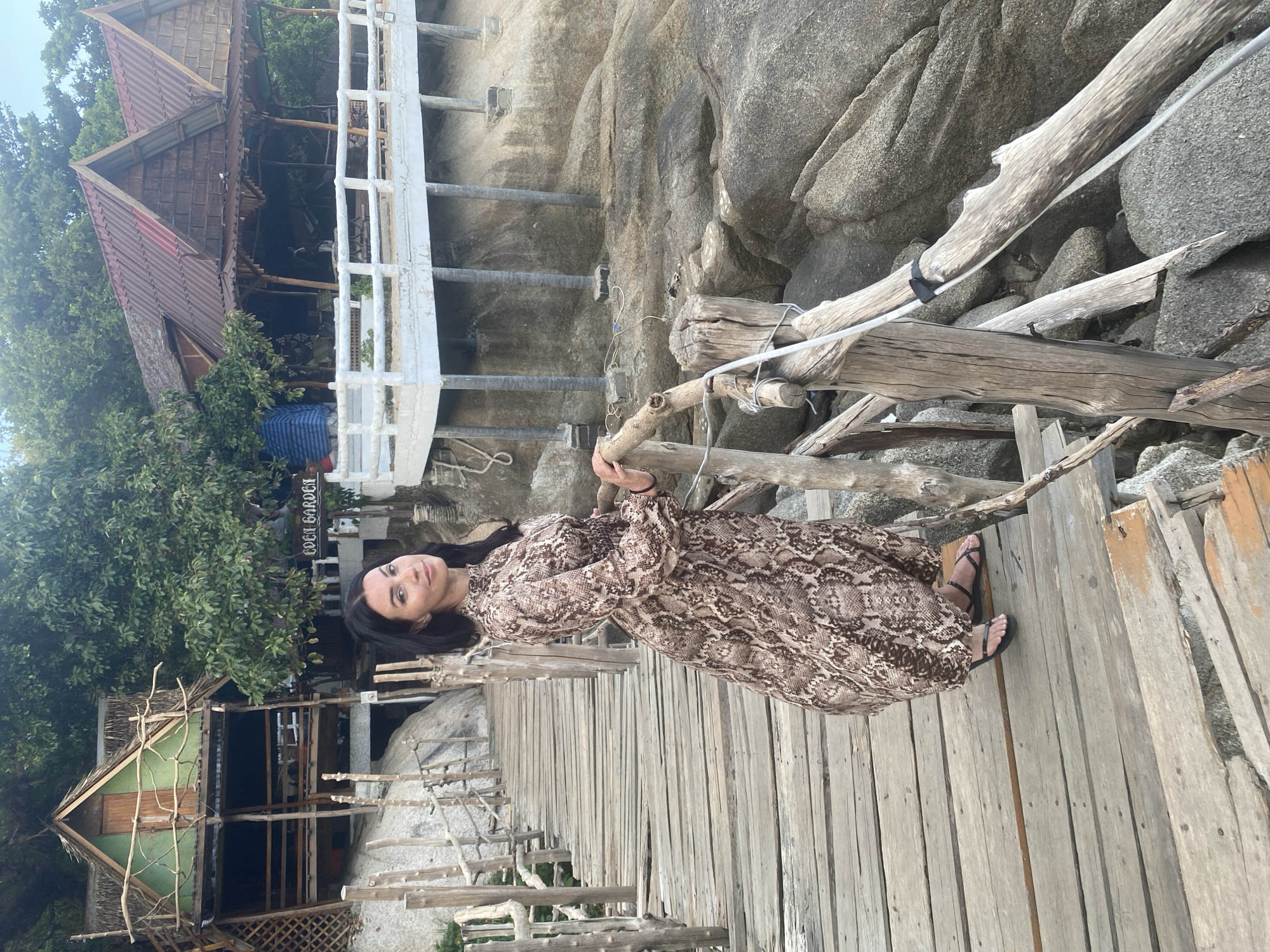 A woman stands on a wooden boardwalk, holding onto a wooden railing. She wears a long, brown patterned dress and black sandals. Behind her, there is a rocky cliff face and a white, elevated walkway. To the left, there are trees and buildings, including one with a sign that reads "EDEN GARDEN". The location is Ban Haad Rin, Thailand.