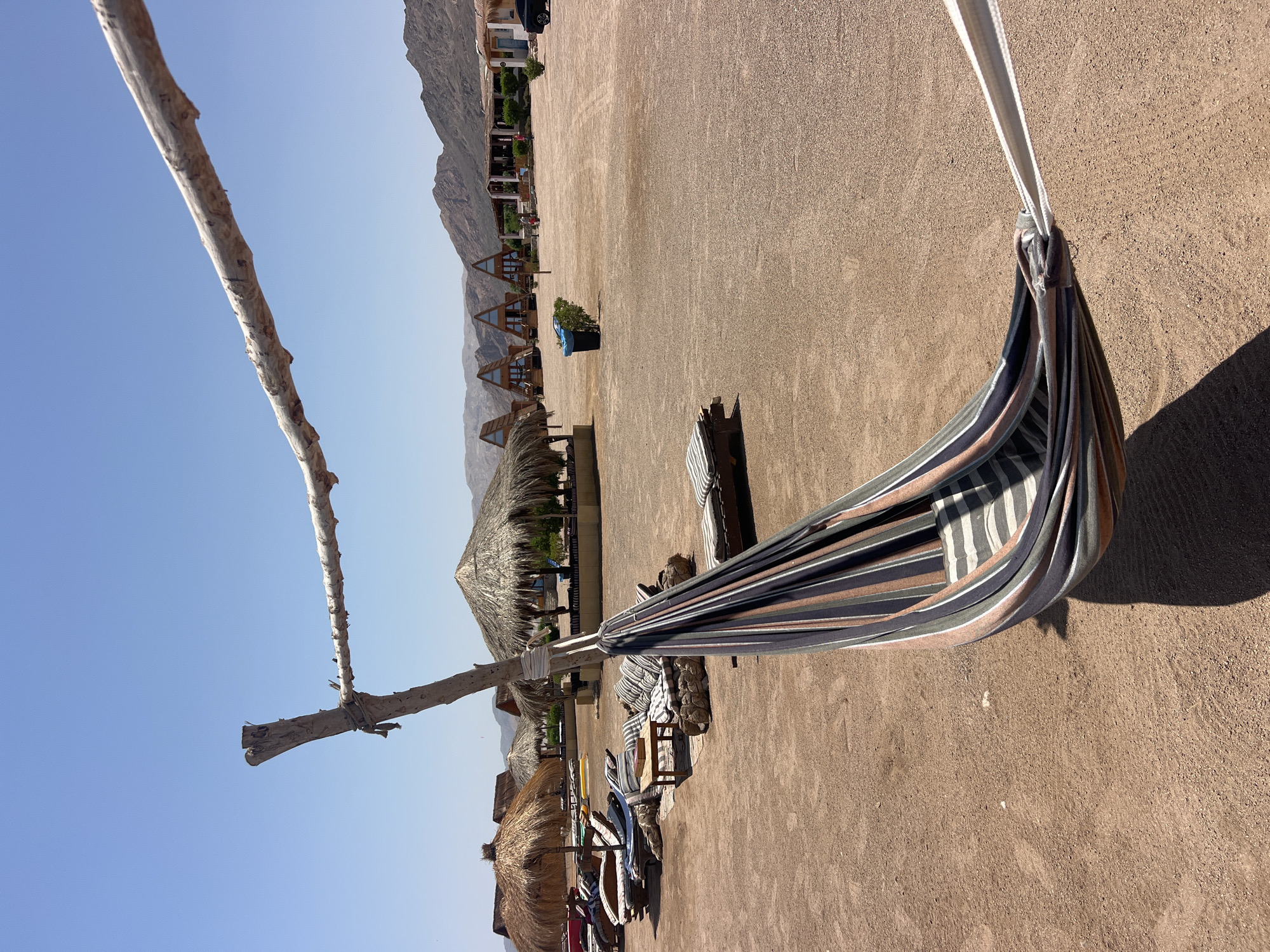A wide-angle, eye-level shot captures a desert resort scene under a clear blue sky in Nuweiba, Egypt. In the foreground, a colorful striped hammock hangs suspended from a rustic wooden frame. The hammock is made of fabric with stripes of gray, white, pink, and dark blue. To the left of the hammock, several lounge chairs and a low wooden table with cushioned seating are arranged on the sandy ground. Beyond these seating areas, thatched-roof umbrellas provide shade over more lounge furniture. In the midground, a cluster of triangular wooden cabins with blue accents are visible. Further back, a long, low-lying building with a white facade and windows is present, and a dark-colored SUV is parked to its right. The entire area is characterized by a sandy, arid landscape with mountains in the distant background. The overall impression is one of a relaxed, desert retreat.