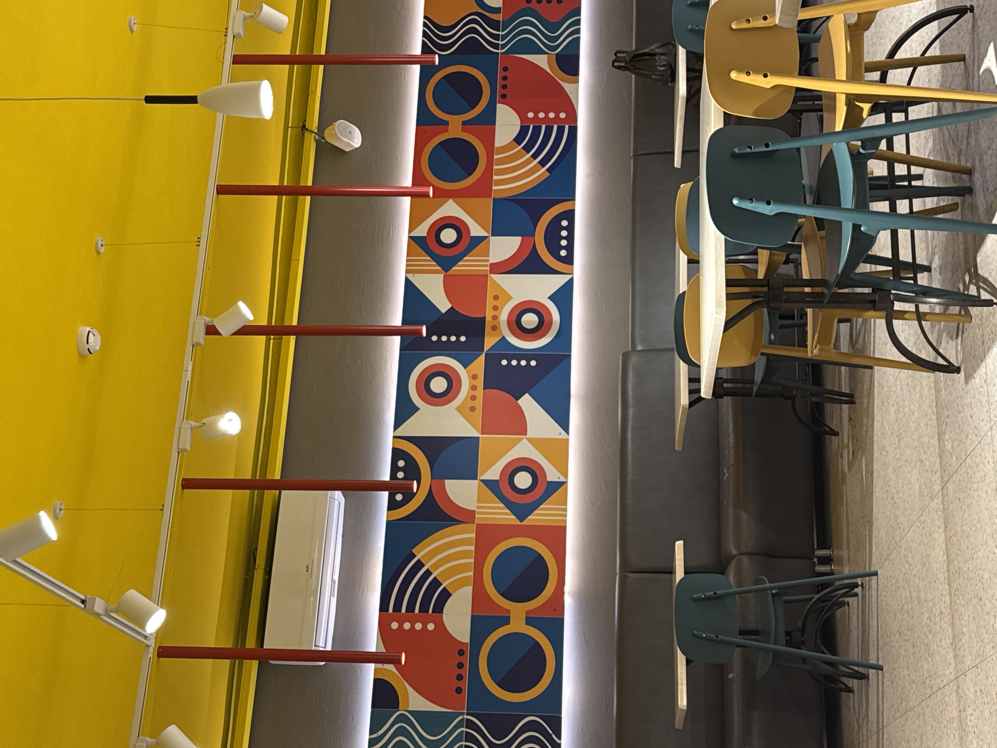 A photograph taken from above, possibly from the ceiling. It depicts a modern interior, presumably a cafe or restaurant. The walls are primarily yellow, with a strip of patterned tiles in shades of blue, orange, white, and red running down the center. Red metal bars extend across the yellow wall. A track lighting system with white fixtures is mounted on the yellow wall. Seating includes a long, dark grey bench or banquette. Several chairs are stacked upside down on the table, featuring a mix of yellow and blue seats. The floor is pale and appears to be tiled. The overall impression is of a clean, modern space that is currently closed, or the chairs are stacked for cleaning. The location in the city of Berezniki, Russia, cannot be determined from the image.