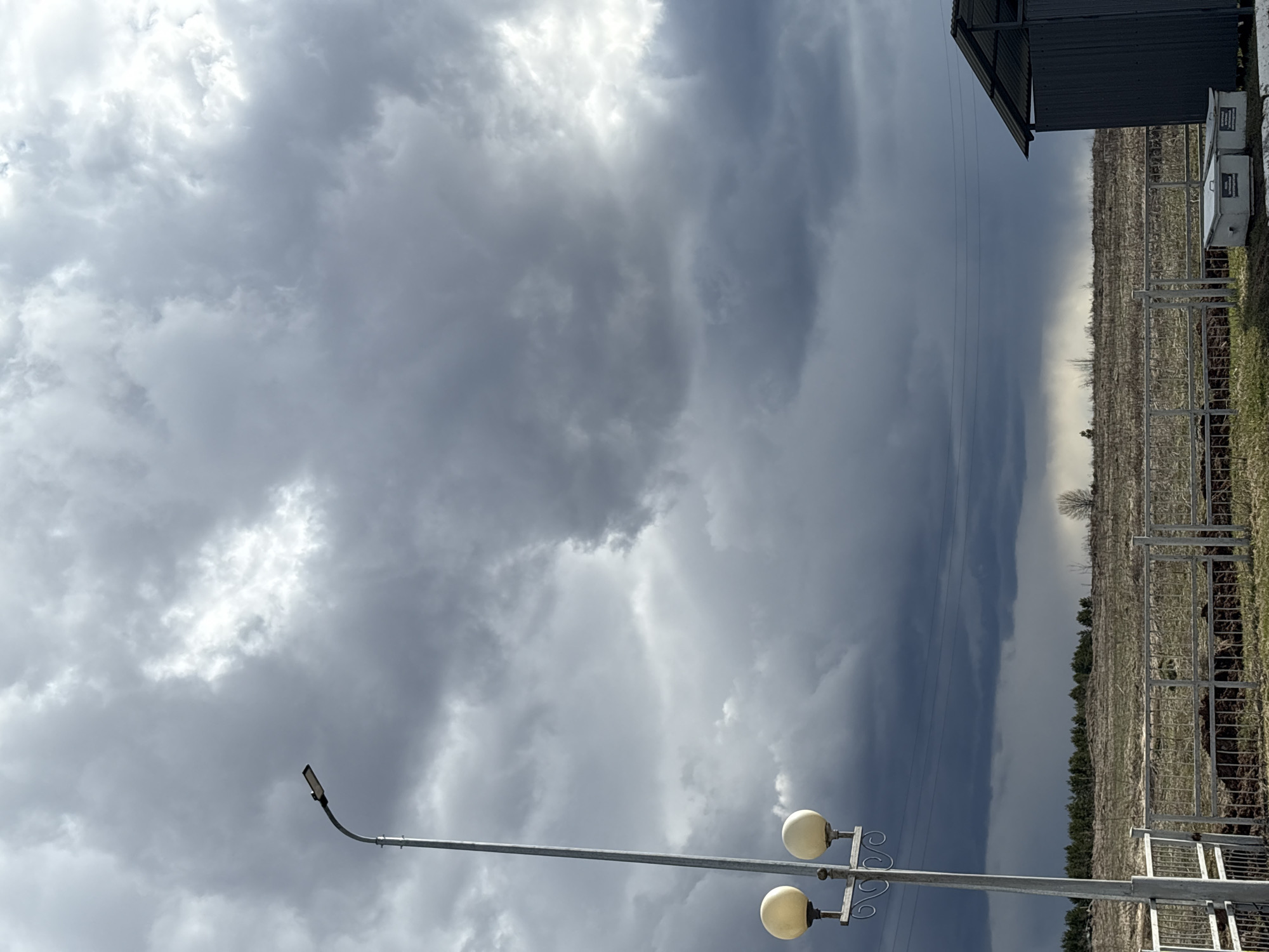The image, originally oriented vertically but depicting a horizontal landscape, presents a dramatic, heavily overcast sky above a rural scene in Alnashi, Russia. The sky is dominated by dense, dark grey storm clouds, particularly pronounced on the right and along the horizon where they form a striking, almost wall-like boundary. Brighter, white-tinged clouds are visible towards the upper left, indicating where diffused sunlight might be penetrating.
In the mid-ground, a barren cultivated field or vineyard stretches across the frame, characterized by numerous vertical poles or trellises, suggesting a non-growing season. A metallic fence runs across the foreground, separating it from the field. A distant line of trees is visible beneath the dark cloud bank.
Two streetlights are present. To the upper left, a modern, single-head LED-style lamp is mounted on a curved pole. In the lower right, an older, ornamental streetlight features two spherical globes and decorative scrollwork. Overhead power lines are visible horizontally through the dark clouds.
A structure with a dark, corrugated metal roof is partially seen in the far upper right, adjacent to two white, rectangular box-like units (likely equipment or storage) with illegible labels. The scene captures a cloudy, possibly stormy daytime, conveying an atmospheric sense of impending weather. No human activity is visible.