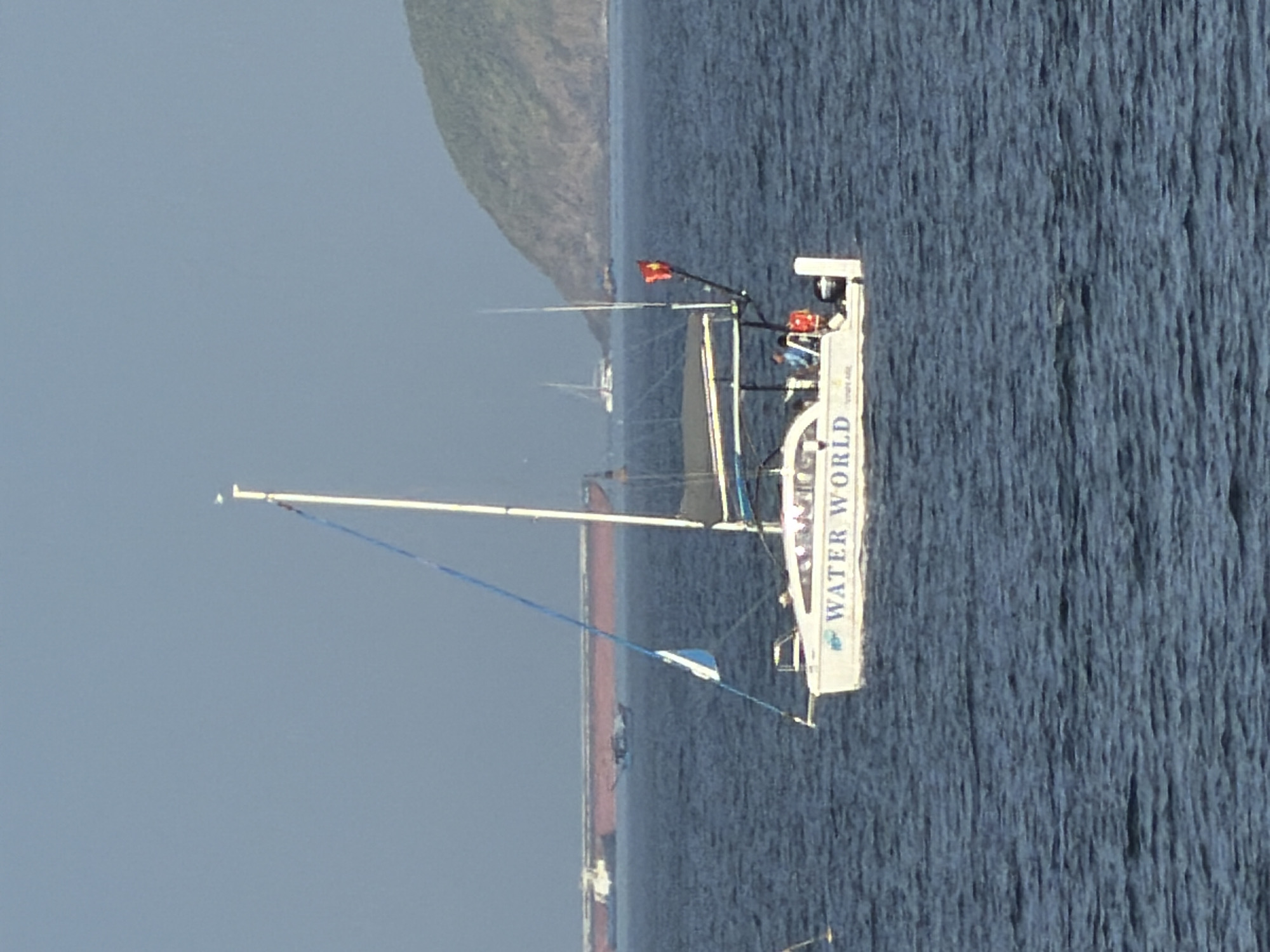 A white sailboat with "WATER WORLD" written on its side is sailing on a blue sea under a clear sky. The boat has a white hull, a mast, and a blue sail. Two people are visible on the boat, one wearing a blue shirt and the other a red life vest. A Vietnamese flag is flying from a pole on the boat. In the background, a large cargo ship and a distant island are visible. The water is calm with small ripples. The scene appears to be set in a coastal area of Nha Trang, Vietnam, given the presence of the Vietnamese flag and the characteristic island landscape.