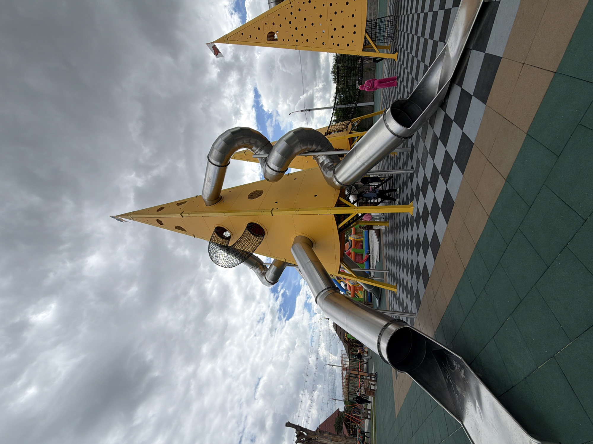 A low-angle, wide shot captures a bright yellow, conical playground structure designed to resemble a piece of cheese, situated in a park in Velykyi Khodachkiv, Ukraine. The structure features several metallic, spiraling slides emerging from its sides, with one slide prominently extending towards the foreground. The playground is surfaced with interlocking rubber mats in green, tan, and black and white checkered patterns. In the background, other playground equipment, including a bouncy castle and swings, are visible, along with a few people, some of whom appear to be children. The sky is overcast with dramatic, gray clouds. A flag with a red and white emblem is visible at the apex of a second, similar yellow structure on the right side of the frame.