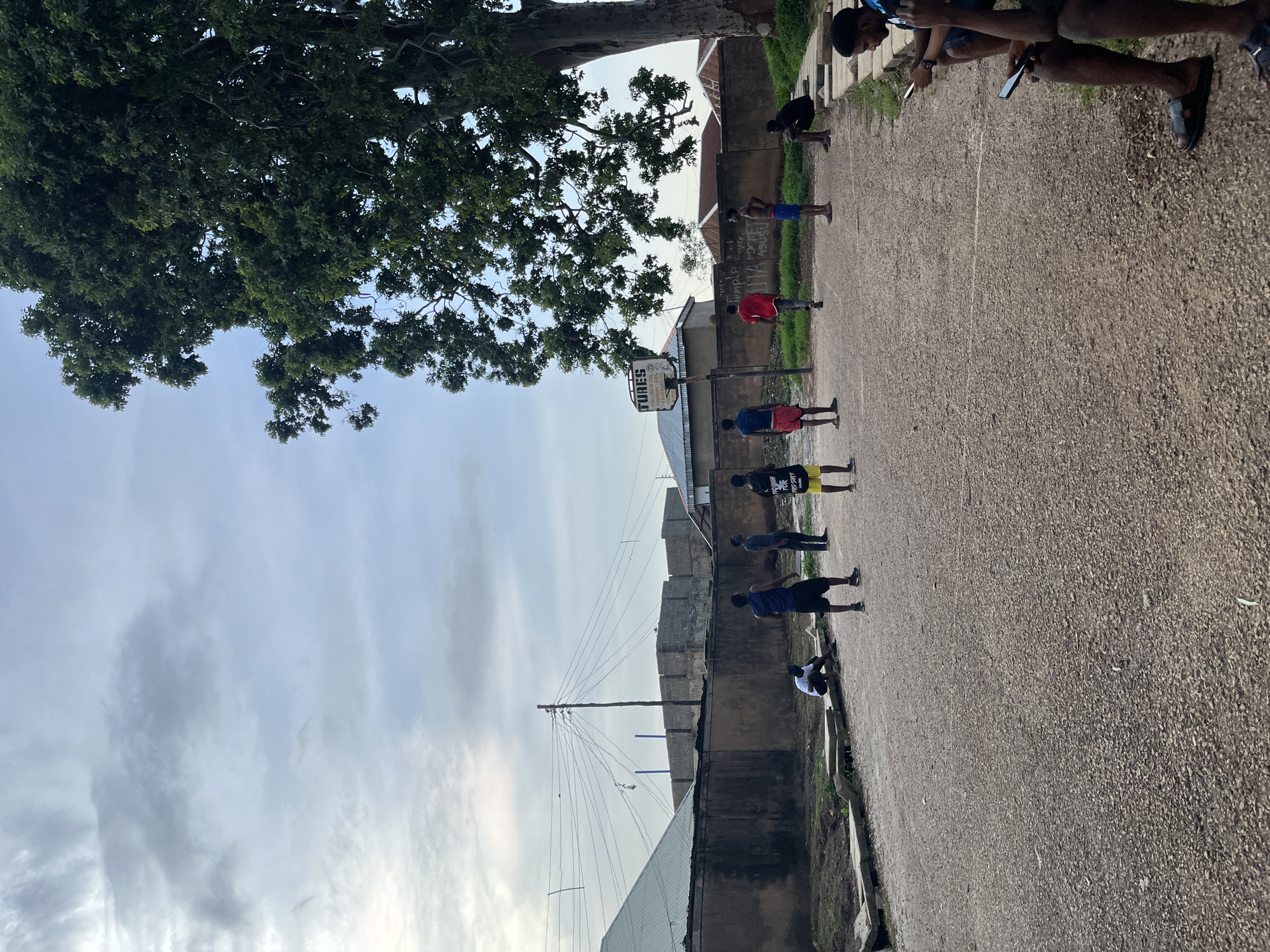 In the city of Jos, Nigeria, on a basketball court with a gravel surface, a group of young men are engaged in various activities under a cloudy sky. A large, leafy tree dominates the right side of the frame, casting shadows. In the background, a concrete wall separates the court from a street lined with houses and a construction site. A basketball hoop with a sign reading "TUNES" is visible. Some men are playing basketball, while others are sitting on the edge of the court, looking at their phones. The overall scene depicts a casual gathering in an urban environment.