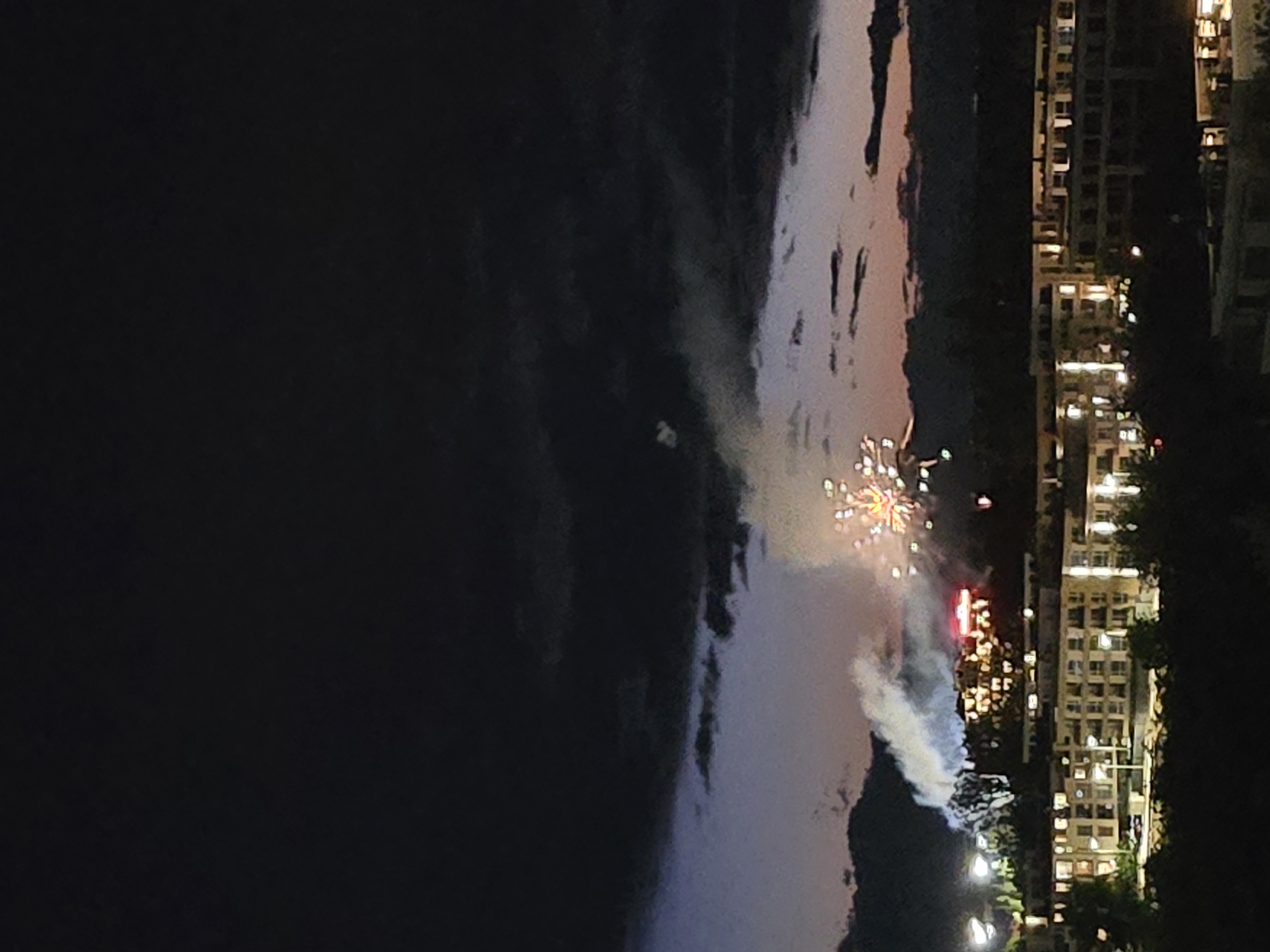 The image depicts fireworks in the city of Unknown, Unknown at night. The fireworks explode in the center of the frame, illuminating smoke and light around them. To the right of the fireworks are tall buildings with numerous lit windows, indicating residential or commercial occupancy. The background is a dark, indistinct body of water or dark open space on the left side of the frame. The general environment appears to be urban, with the buildings suggesting a densely populated area. There are no people visible in the image.