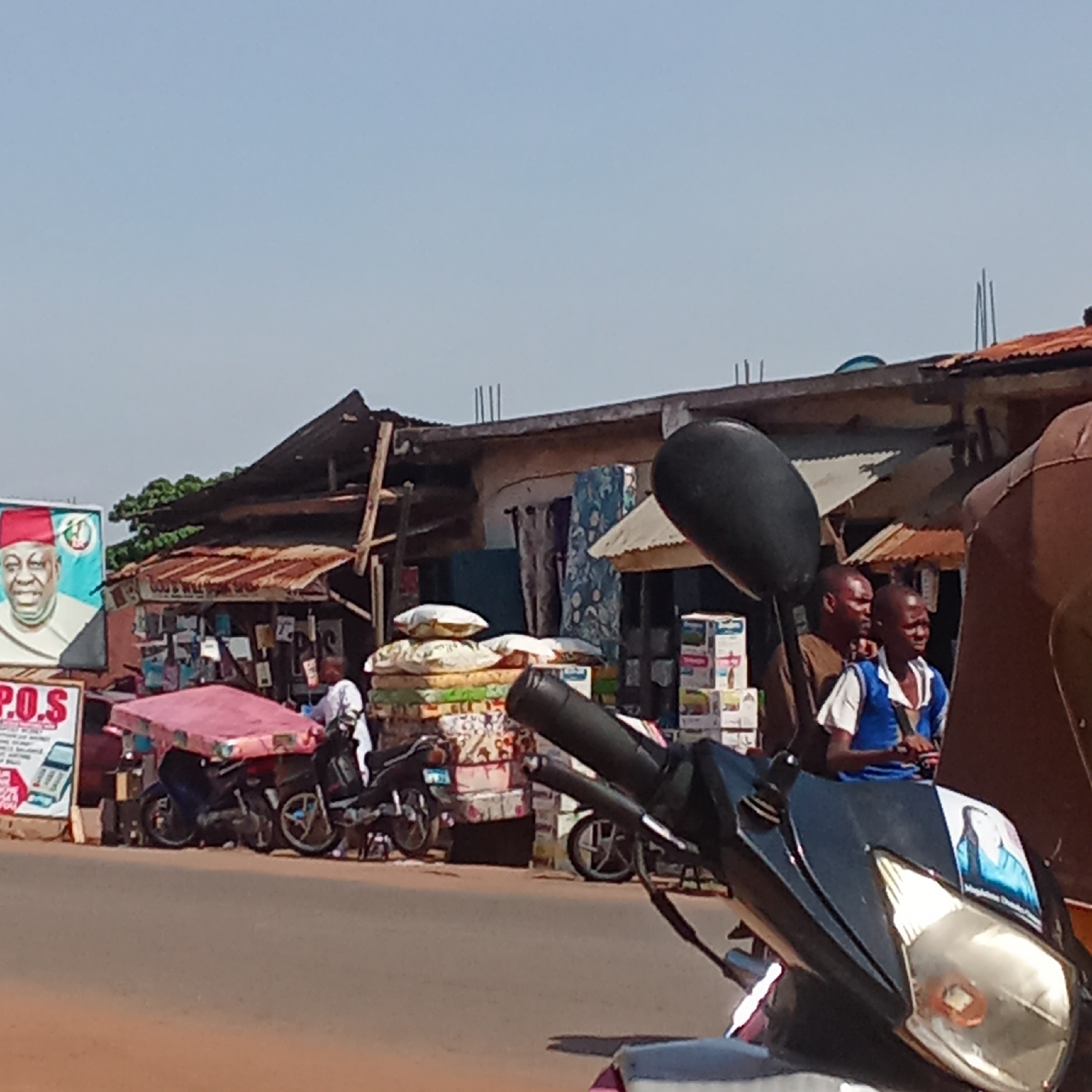 The image depicts a street scene in Doma, Nigeria. Several people are present, including two boys standing near a black motorcycle in the foreground. The motorcycle has a sticker of a woman on its front. Further back, individuals are visible near a roadside market. The market consists of shops or stalls with corrugated iron roofs. Goods, including mattresses and stacked boxes, are displayed. Beside the market, a large poster featuring a man in a red hat is visible. The road in the foreground is asphalt. The sky is clear and blue.