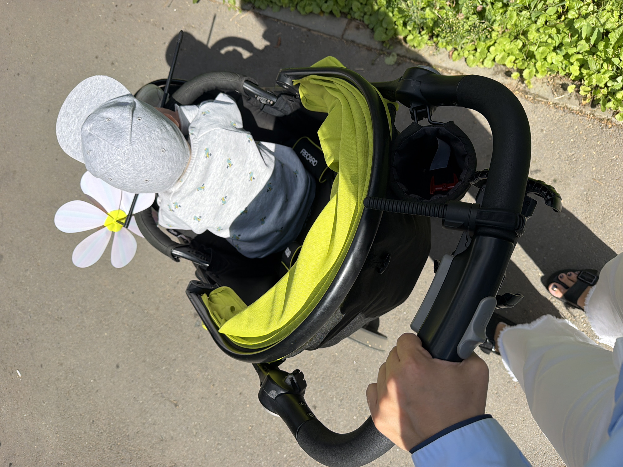 A high-angle, over-the-shoulder shot captures a person pushing a baby in a stroller on a paved surface, likely a sidewalk or path, in Votkinsk, Russia. The baby, facing away from the camera, is seated in a black stroller with a bright lime-green fabric interior and wears a grey cap and a light grey onesie with a repeating pattern of small blue birds. A large, white, pinwheel-like decoration with a yellow center is attached to the stroller. The person pushing the stroller is wearing a light blue shirt and white pants, and black sandals on their feet. The ground is asphalt, and to the right, a strip of green vegetation lines the path. The sun casts strong shadows on the ground, indicating a bright, sunny day.