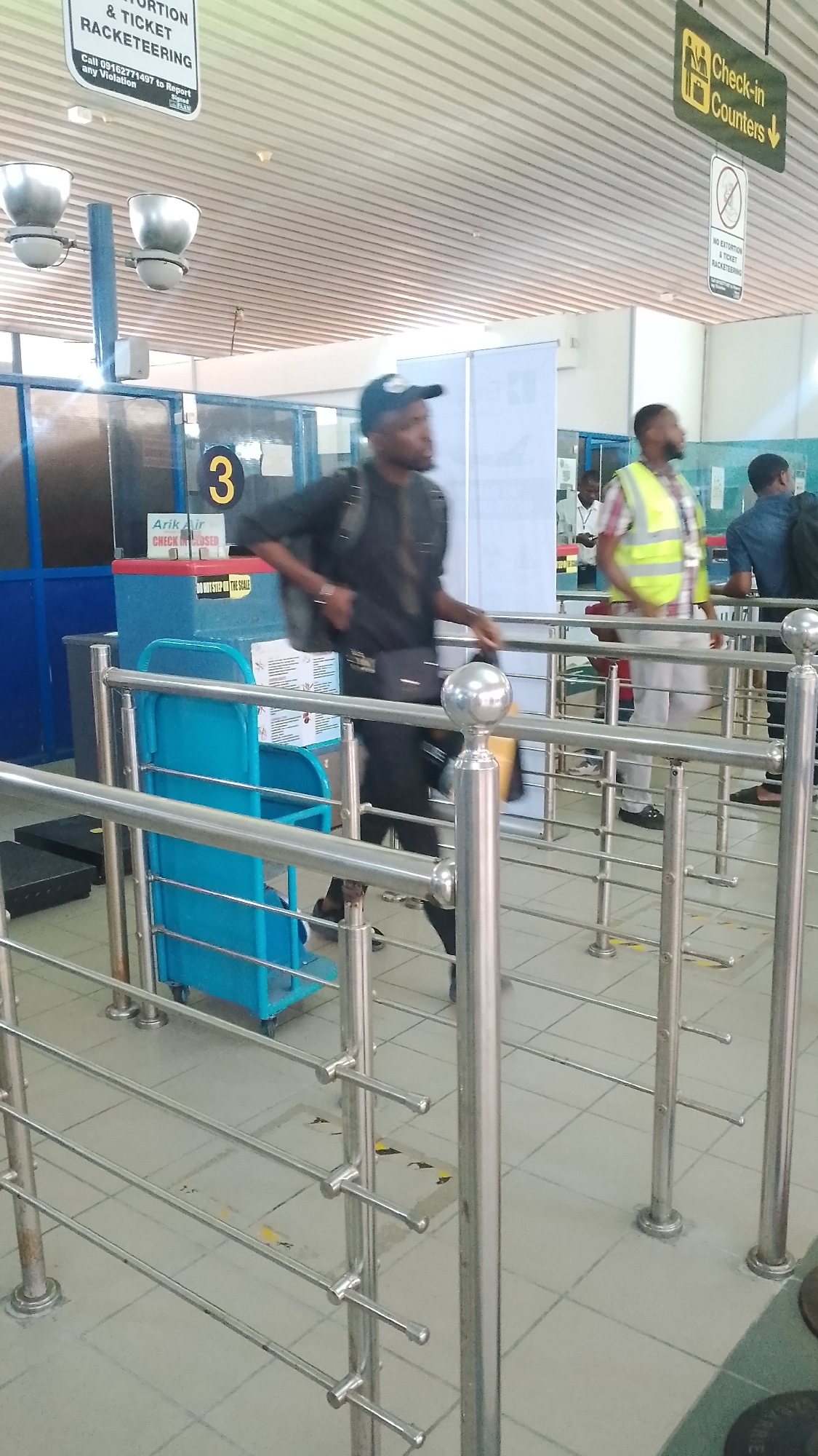 The image shows the interior of an airport terminal, likely in Nigeria given the signage in the background. Several people are visible, including one man in a black shirt and cap walking through metal queue barriers. Behind him, another man wears a yellow safety vest. Check-in counters are visible in the background with signage indicating "Check-in Counters." Other signs advise against extortion and ticket racketeering, providing a phone number to report violations. An "Arik Air" check-in counter is marked as "Closed". There is a blue luggage cart near the queue barriers. The location in Orerokpe, Nigeria is not confirmed, but inferred from context.