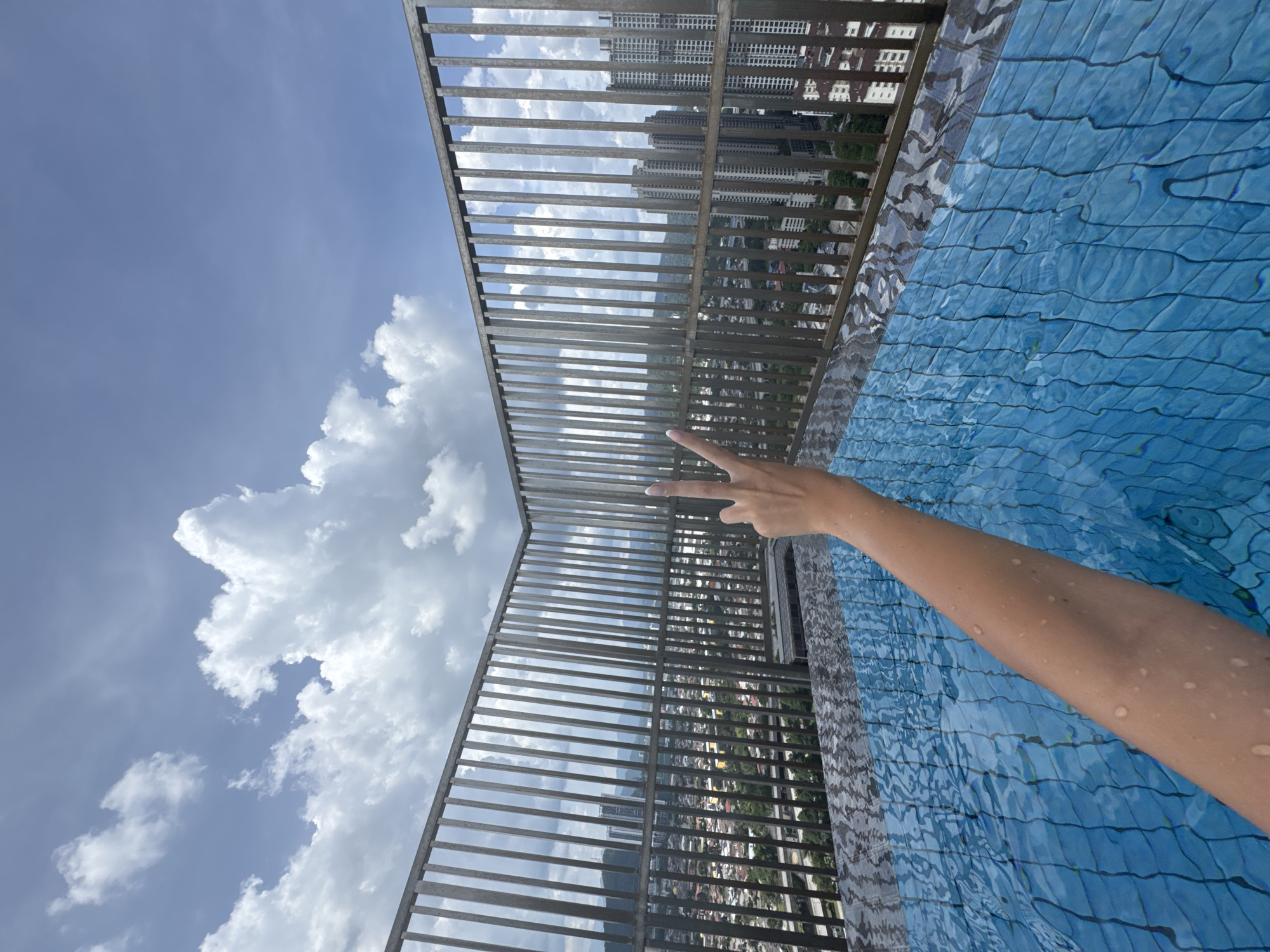 This image captures a vibrant overhead view from a high-rise location, likely a rooftop or elevated pool, in Ampang Jaya Municipal Council, Malaysia. The main subject is a person's wet arm and hand, extending into the frame from the lower right and making a "peace" or "V" sign.
To the right, the bright blue water of a swimming pool is visible, featuring a distinct patterned tile bottom and ripples on its surface, suggesting movement or an infinity edge. A sturdy metal railing, composed of vertical slats, runs across the middle of the frame, separating the pool area from the expansive background.
Beyond the railing, a dense cityscape unfolds under a clear, bright blue sky dotted with fluffy white clouds. Numerous high-rise buildings are visible, indicating an urban environment. The scene is bathed in sunlight, suggesting a clear and sunny daytime. There is no visible text in the image.