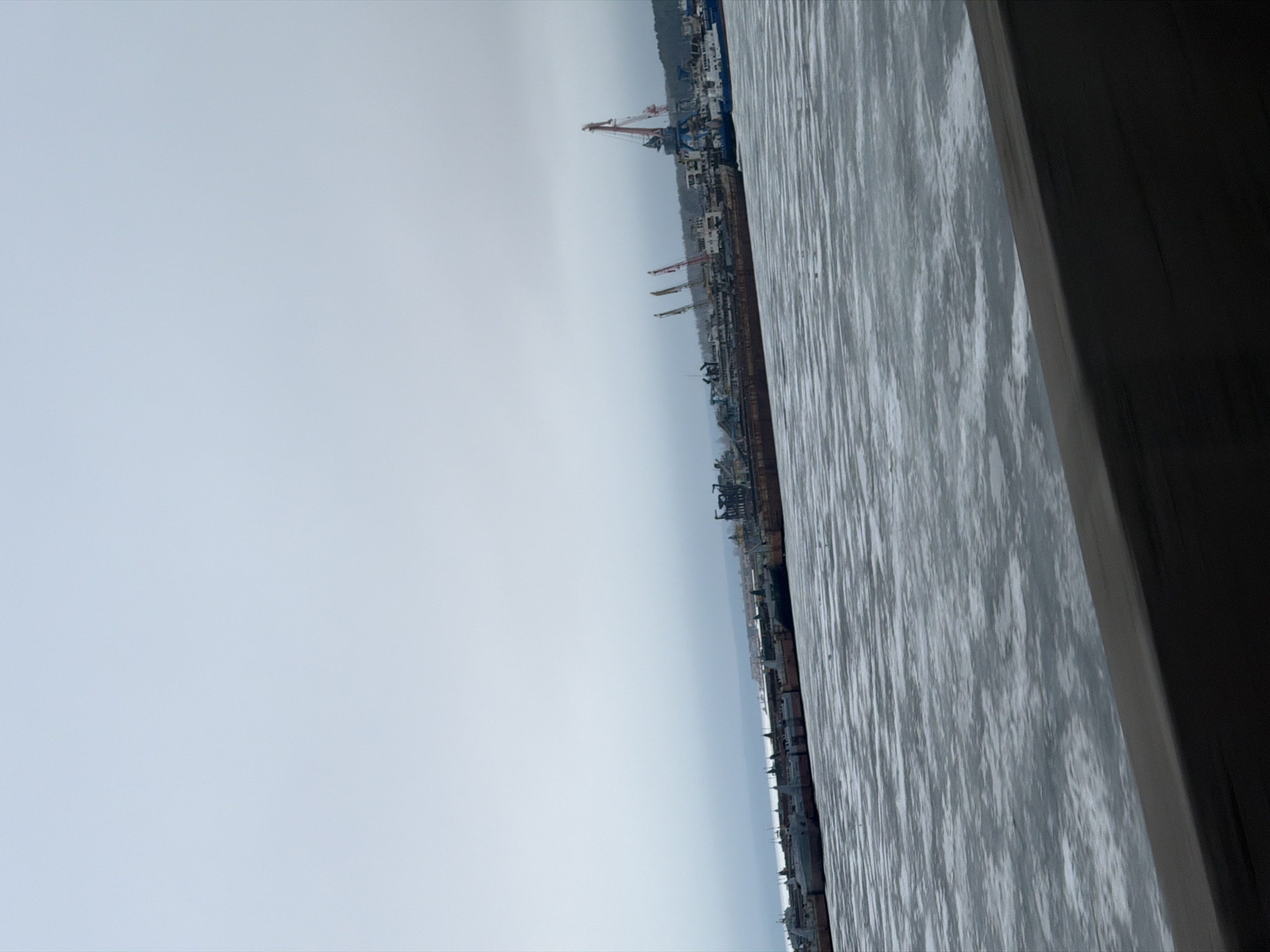 The image shows a waterfront view from an elevated position. The water appears textured, possibly due to waves or ice. In the distance are industrial structures including cranes and buildings. To the right, a dark, angled structure is visible, potentially part of a building or viewing platform. The sky is overcast, and the overall scene has a gray tone. The location is Chaikovsky, Russia.