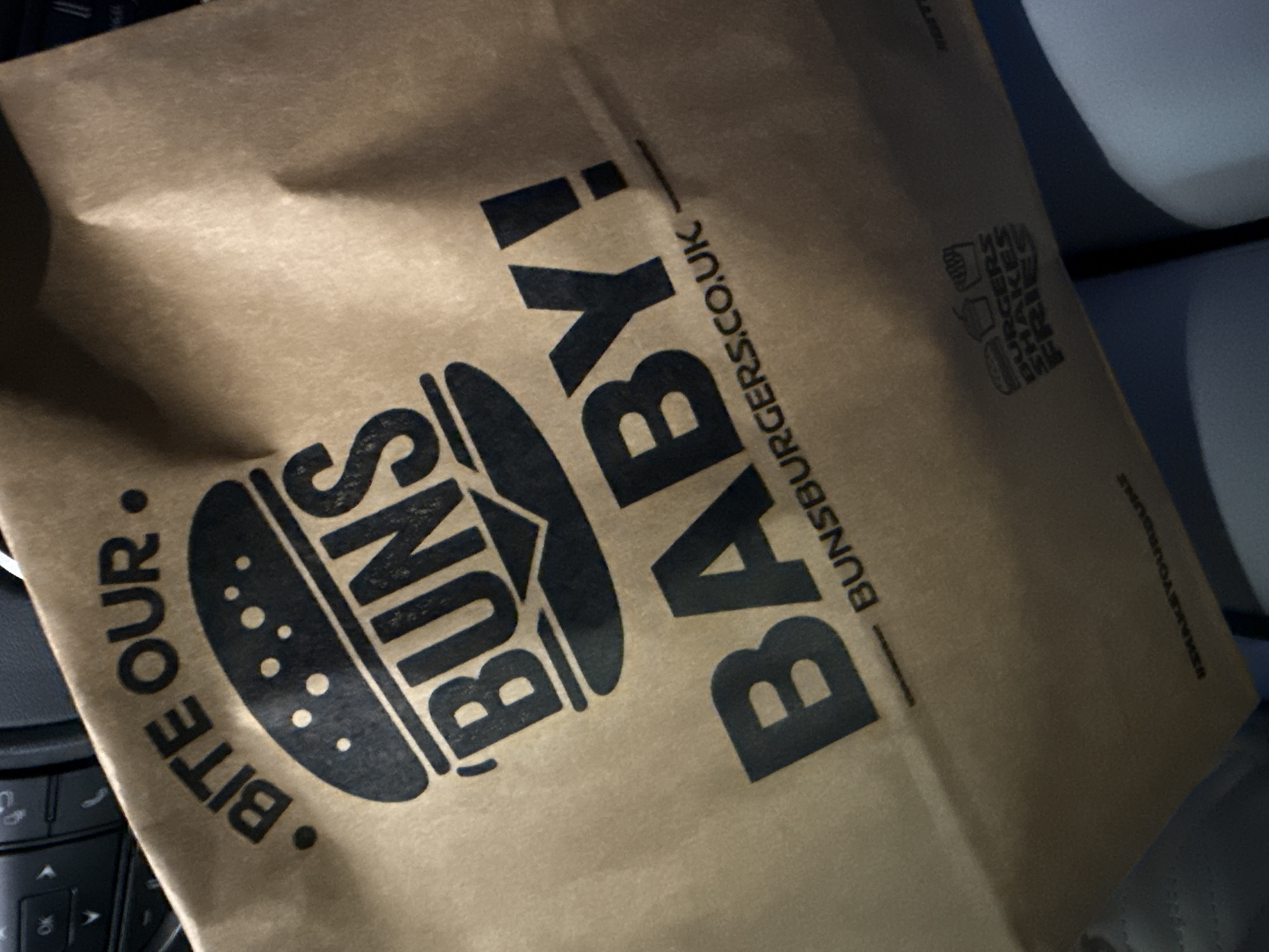 A tan paper bag from "BUNS BURGERS.CO.UK BABY!" is shown, advertising burgers. The bag also contains the words "BITE OUR". The words "BURGERS SHAKES FRIES" are on the bag as well. The bag appears to be sitting on a car seat near the center console. The location is most likely Nottingham, United Kingdom, due to the domain name on the bag.