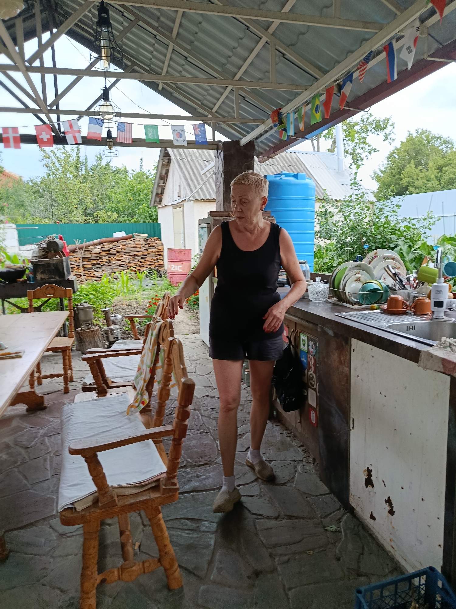 An elderly woman with short, blonde hair and fair skin, wearing a black tank top and black shorts, stands next to an outdoor kitchen counter. She has her right hand resting on a wooden chair with a white cushion. The kitchen counter is cluttered with dishes, some in a sink, and a blue water tank is visible behind her. In the background, a wooden table and chairs are set up, and a pile of firewood is stacked against a building. A string of international flags hangs overhead. The scene appears to be an outdoor patio or backyard area in a rural setting.