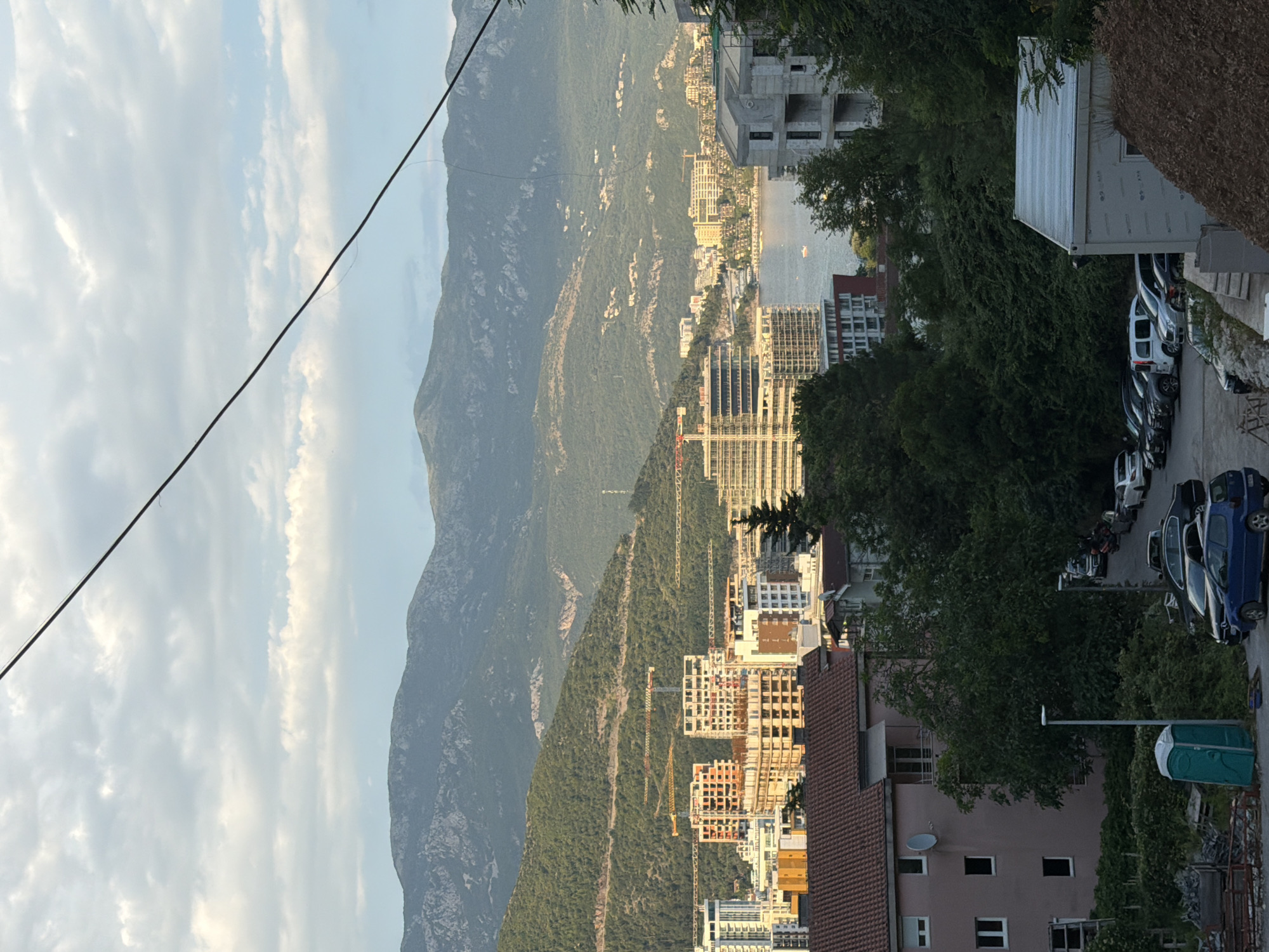 The image depicts a panoramic view of Budva, Montenegro, as seen from an elevated perspective. In the foreground, several parked cars are visible on a paved area adjacent to lush green foliage. A portable toilet is situated to the left of the parked cars, near a pink building with multiple windows. Further in the mid-ground, a dense collection of trees forms a green barrier, separating the foreground from a developing urban landscape. This urban area is characterized by numerous multi-story buildings, many of which appear to be under construction, indicated by visible cranes. A body of water, likely the Adriatic Sea, can be seen behind the buildings, with a small boat on its surface. The background is dominated by a majestic mountain range, its slopes covered in greenery. The sky above is partly cloudy, with soft sunlight illuminating the scene, suggesting it is either early morning or late afternoon. The overall impression is one of a coastal city with ongoing development, set against a dramatic natural backdrop.