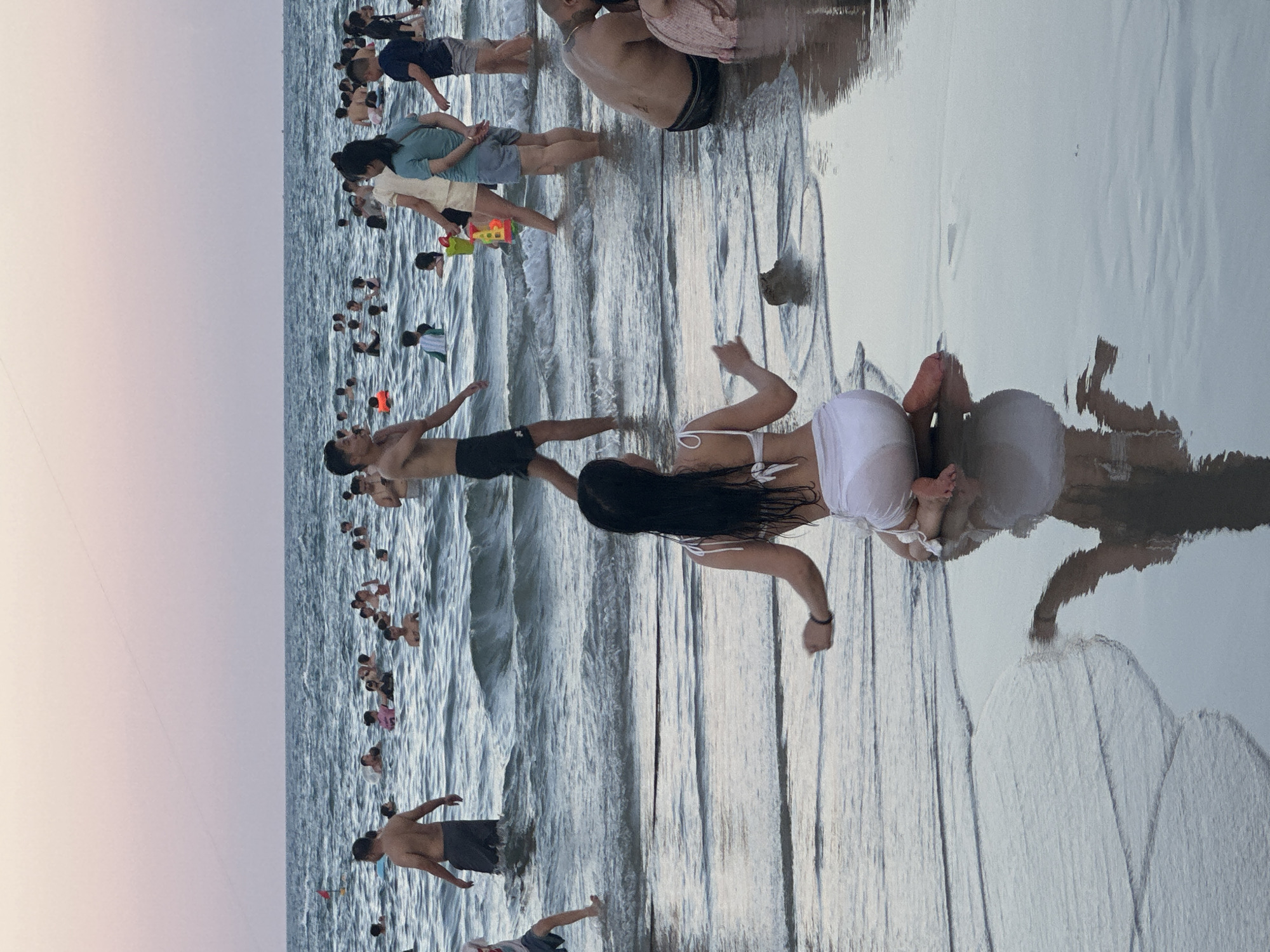 The image depicts a crowded beach scene in Đà Nẵng, Vietnam, likely during the late afternoon or early evening given the soft, diffused light. The sky is a pale pink-purple gradient, suggesting either sunrise or sunset. The ocean is calm with gentle waves lapping at the shore, and the wet sand reflects the sky and the people on the beach.
Numerous individuals are present, engaged in various beach activities. Many are wading or standing in the shallow water, some appearing to be swimming or playing. A group of people are clustered further out in the water, while others are closer to the shore. In the foreground, two individuals are more prominently featured. A man is standing with his back to the camera, facing the ocean, and a woman is lying on her stomach in the shallow water, her body angled towards the viewer. She is wearing a white bikini and her long dark hair is spread out. The man appears to be wearing dark shorts, and the woman is wearing a white bikini bottom and a white top.
Other notable details include a red float or buoy in the water, and a child's colorful toy shovel and bucket visible on the sand to the left of the prominent figures. The overall atmosphere seems relaxed and social, with people enjoying the beach. The water is clear enough to see the sand beneath it, and the reflections on the wet sand create a mirror-like effect. There is no visible text in the image.