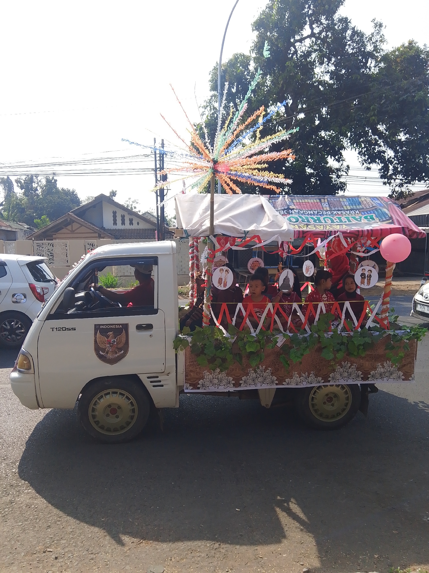 A white pickup truck is decorated as a parade float in Kalinyamatan, Indonesia. The truck has a canopy over the back, adorned with red and white streamers and circular ornaments featuring images of people and cultural landmarks. Children in red shirts are seated on the float. A pink balloon is tied to the right side of the canopy. The truck features an "INDONESIA" emblem on its side. A man is driving the truck, wearing a red shirt and a cap. The background shows trees, houses, and a utility pole with colorful decorations. The scene appears to be taking place on a sunny day.