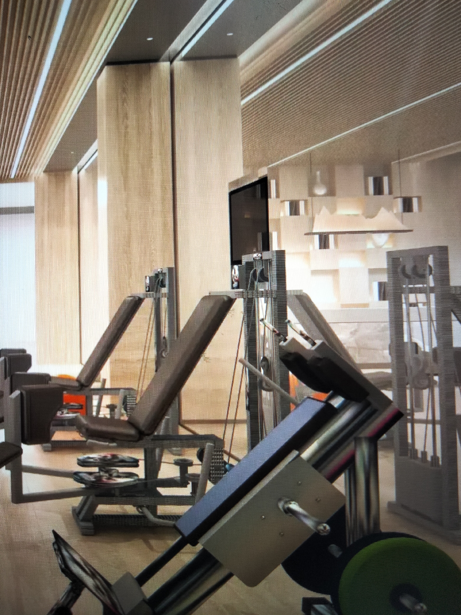 The image shows an indoor gym with several weight machines. The gym has a modern design with light wood paneling on the walls and ceiling. In the foreground, there is a weight machine with a dark gray seat and chrome-plated levers. A large green weight plate is visible at the bottom right of the image. In the background, there are more weight machines, including leg press machines with brown padded seats. A television screen is mounted on a pillar. The floor appears to be made of wood or a similar material. The overall impression is one of a clean, well-equipped, and contemporary fitness facility. The location is specified as Payagaboh, Indonesia.