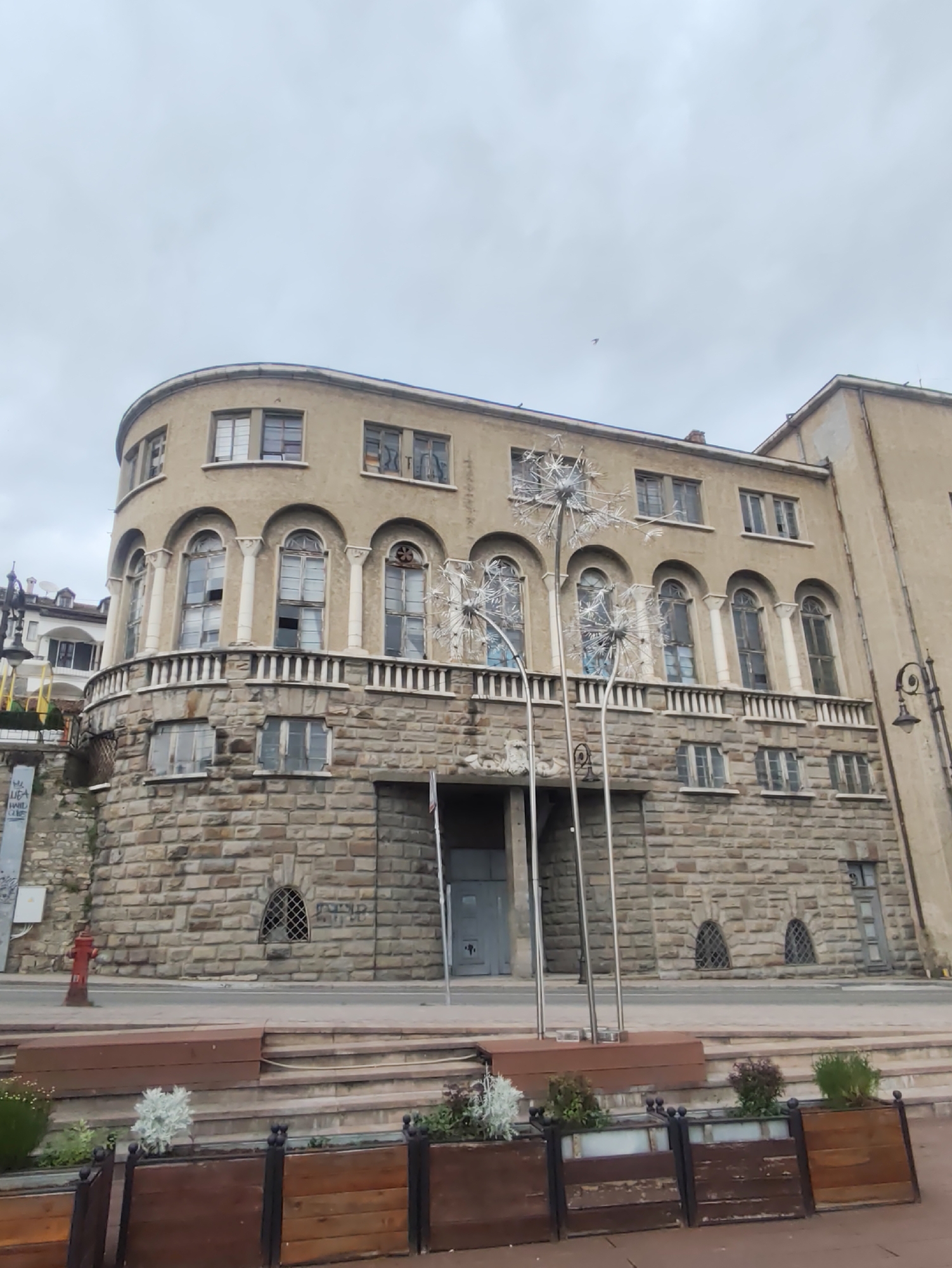 The image shows a building in Veliko Tarnovo, Bulgaria. The building has three stories, with the bottom story built of rough-cut stone blocks and the upper stories made of smooth stucco. The building's architecture is characterized by arched windows and a curved facade. In front of the building are decorative metal sculptures resembling dandelions. There are wooden planters with plants in front of a staircase leading up to the building. A red fire hydrant is visible to the left of the building. The sky is overcast and gray.