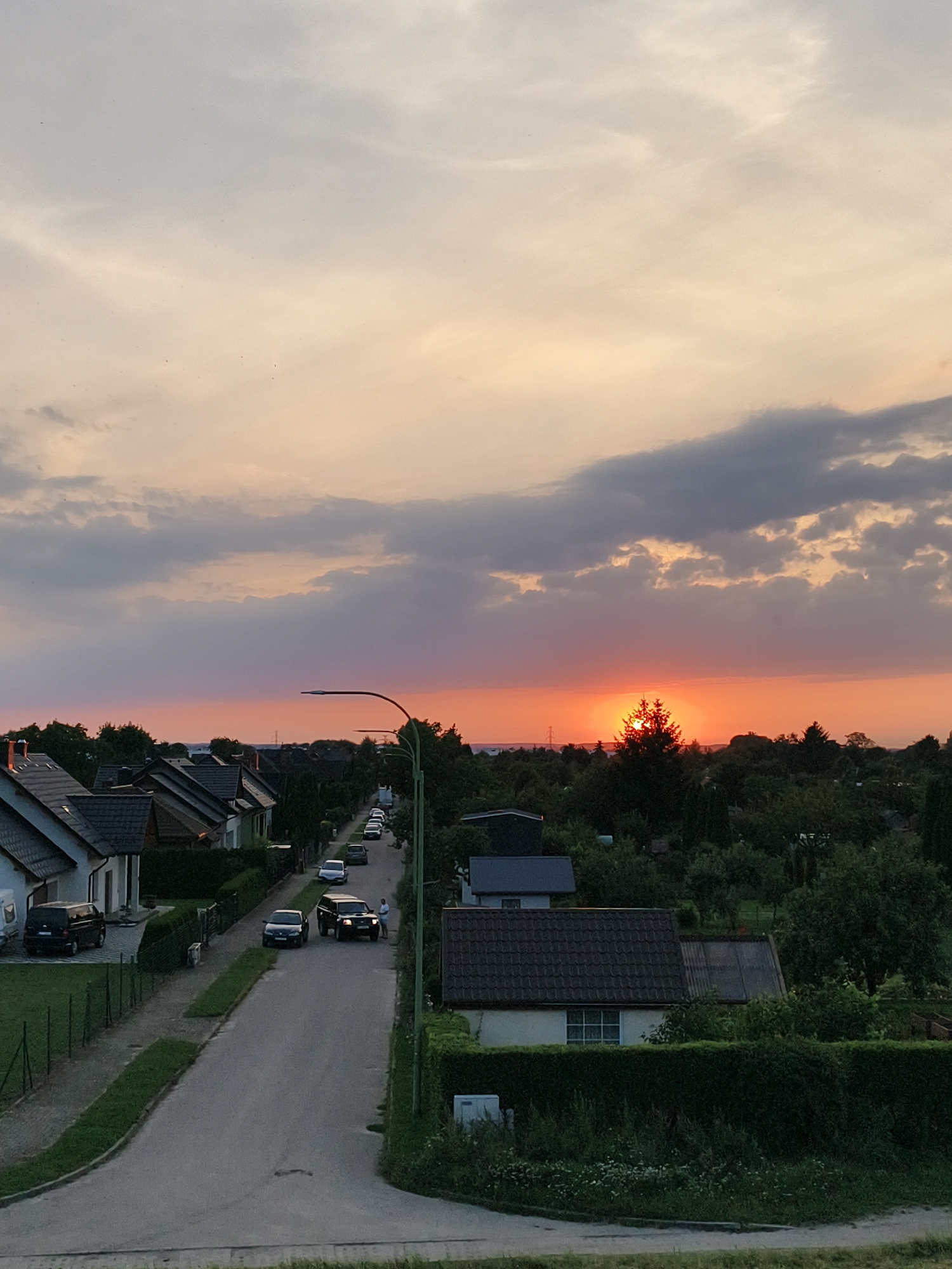 The image depicts a residential street in Tczew, Poland, at sunset. The sky is filled with a gradient of colors, from pale yellow at the top to vibrant orange and pink near the horizon, with scattered clouds. The sun is partially obscured by trees in the distance, casting a warm glow over the scene.
On the street, several cars are visible, including a black van parked on the left, a black sedan, and a dark-colored SUV. Two individuals are standing near the SUV. Houses with pitched roofs line both sides of the street, with well-maintained hedges and gardens. A streetlamp with a modern design is positioned on the right side of the road. The overall atmosphere is serene and tranquil, characteristic of a quiet evening in a suburban neighborhood. The architecture and layout of the houses suggest a typical Polish residential area.