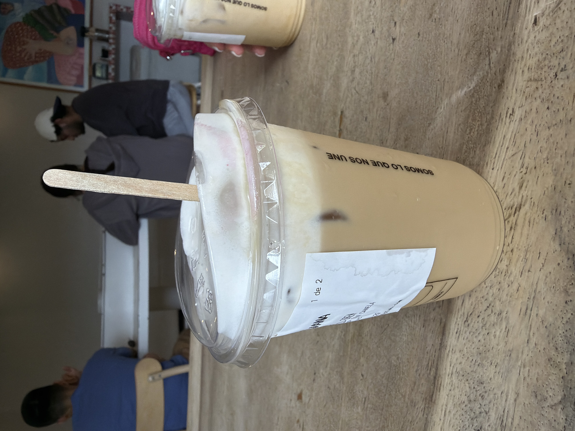 A plastic cup of iced coffee with a wooden stick and foamy topping sits on a wooden table. The cup has a white label with the text "1 de 2" and "MANA" printed on it. The cup is labeled "Somos lo que nos une". Other people are in the background. Based on the label, the location is most likely in the city of Puebla, Mexico.