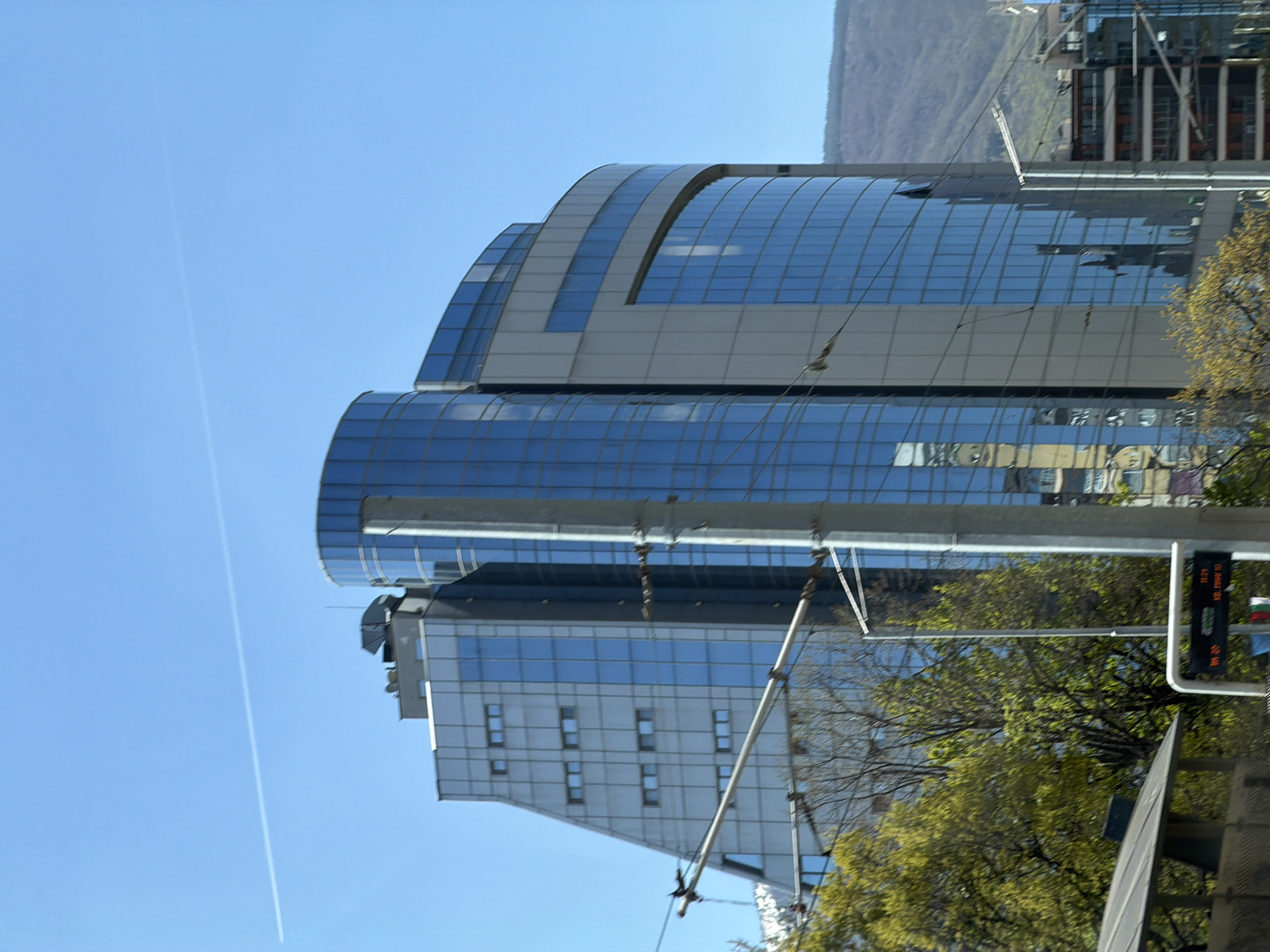 The image captures a modern building with a curved glass facade in Sofia, Bulgaria, under a clear blue sky marked by an airplane contrail. The architecture features repeating patterns of rectangular windows set within a light-colored frame. A mountain range is faintly visible in the background. In the foreground, there are elements of street infrastructure including poles, wires, and traffic signals, partially obscured by green foliage from nearby trees.