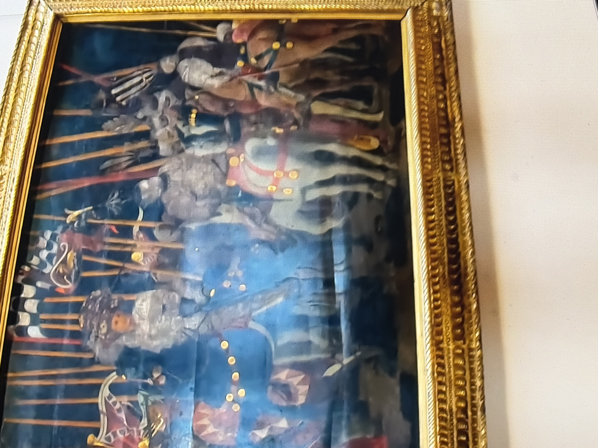 The image depicts a framed oil painting of a procession of armored figures on horseback. The figures are wearing elaborate, ornate armor and elaborate headdresses. The horses are adorned with decorative coverings and golden accents. In the background, a series of upright poles are visible, some of which appear to be topped with flags or banners, one with a checkered pattern. The overall scene suggests a historical or ceremonial event. The painting is presented in a golden, ornate frame. The background appears to be a solid white surface, suggesting the painting is displayed on a wall. No specific location in the city of Unknown, Unknown is identifiable.