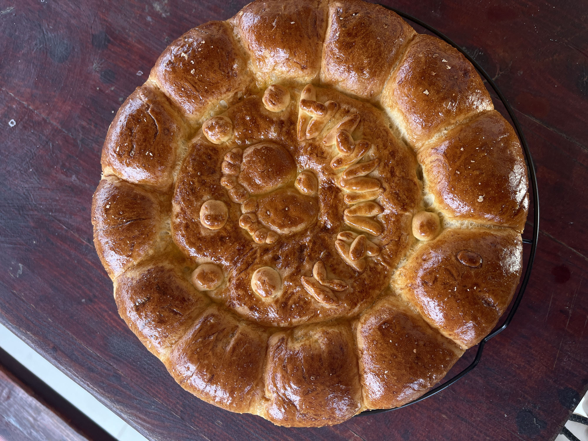 A baked loaf of bread is displayed on a dark wooden surface in the city of Kosten, Bulgaria. The bread is circular with a decorative design. The outer ring of the loaf is composed of a series of smaller, rounded sections. The central portion of the bread features raised patterns of baked dough. The bread has a golden-brown, glazed finish, suggesting it has been brushed with an egg wash or similar substance before baking. The loaf is positioned on a black wire rack. The wooden surface on which the bread is placed has a slightly glossy finish. A white element is visible to the lower left of the frame, potentially a portion of a table or countertop. The lighting in the scene is natural, highlighting the textures of the bread and the wooden surface.