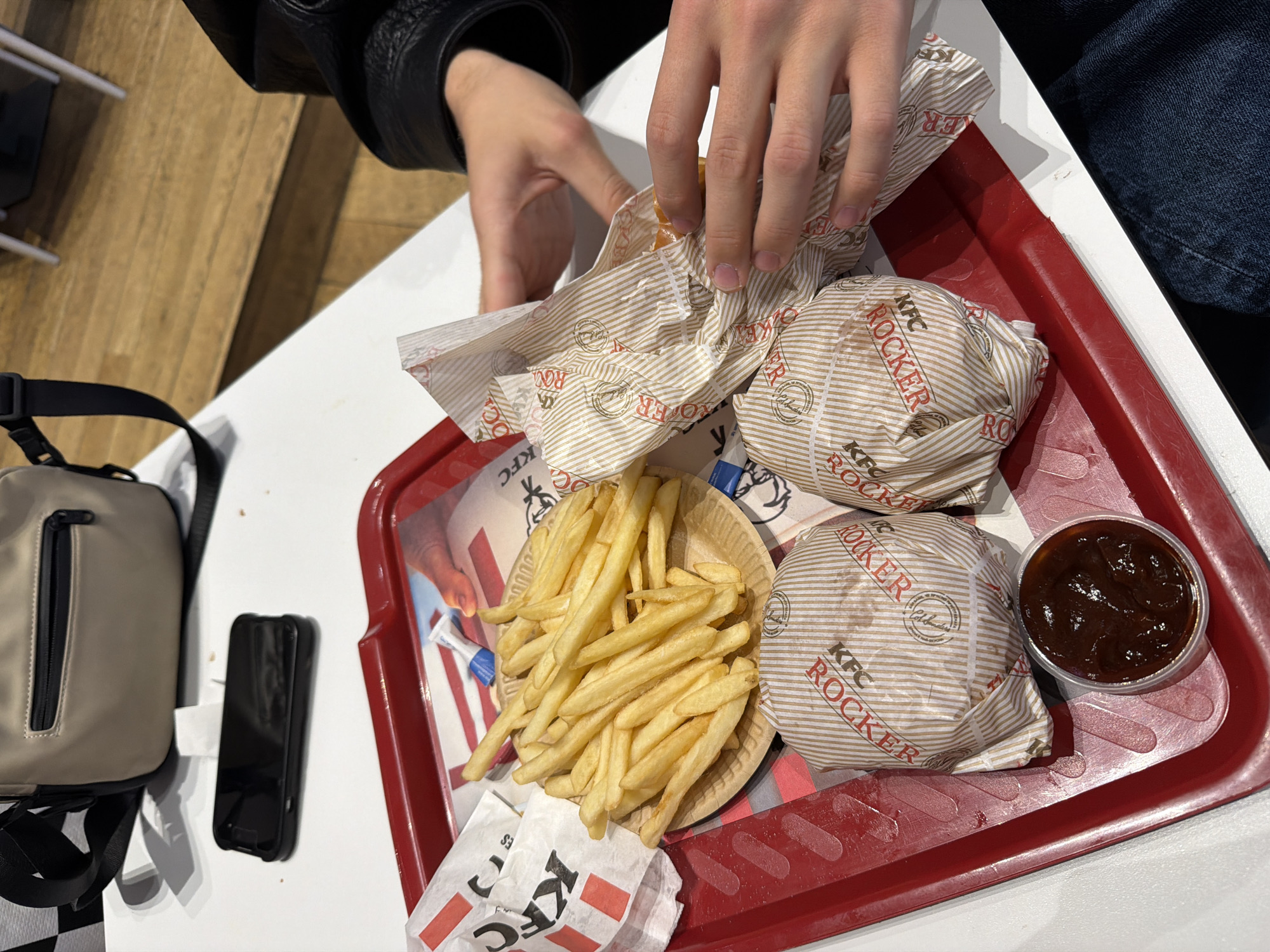 An overhead shot captures a fast-food meal laid out on a white table within what appears to be a KFC restaurant in Sofia, Bulgaria. A person's hands, visible in the upper left of the frame and wearing a black leather jacket, are in the process of unwrapping one of two burgers.
The meal is presented on a rectangular red plastic tray, which shows some signs of wear and scratches. Two burgers, clearly labeled "KFC ROCKER" and featuring the KFC logo on their striped paper wrappers, are central to the tray. Next to the burgers, a generous serving of golden-brown french fries is contained within a round, light brown, wood-patterned bowl. A small, clear plastic cup holds a dark, thick sauce, likely barbecue sauce.
To the left of the tray, a beige or light grey cross-body bag with a black zipper and strap rests on the white table. Adjacent to the bag is a sleek black smartphone. The background shows a wooden floor, suggesting an indoor setting. The image depicts a casual dining experience, with the individual about to consume their meal.