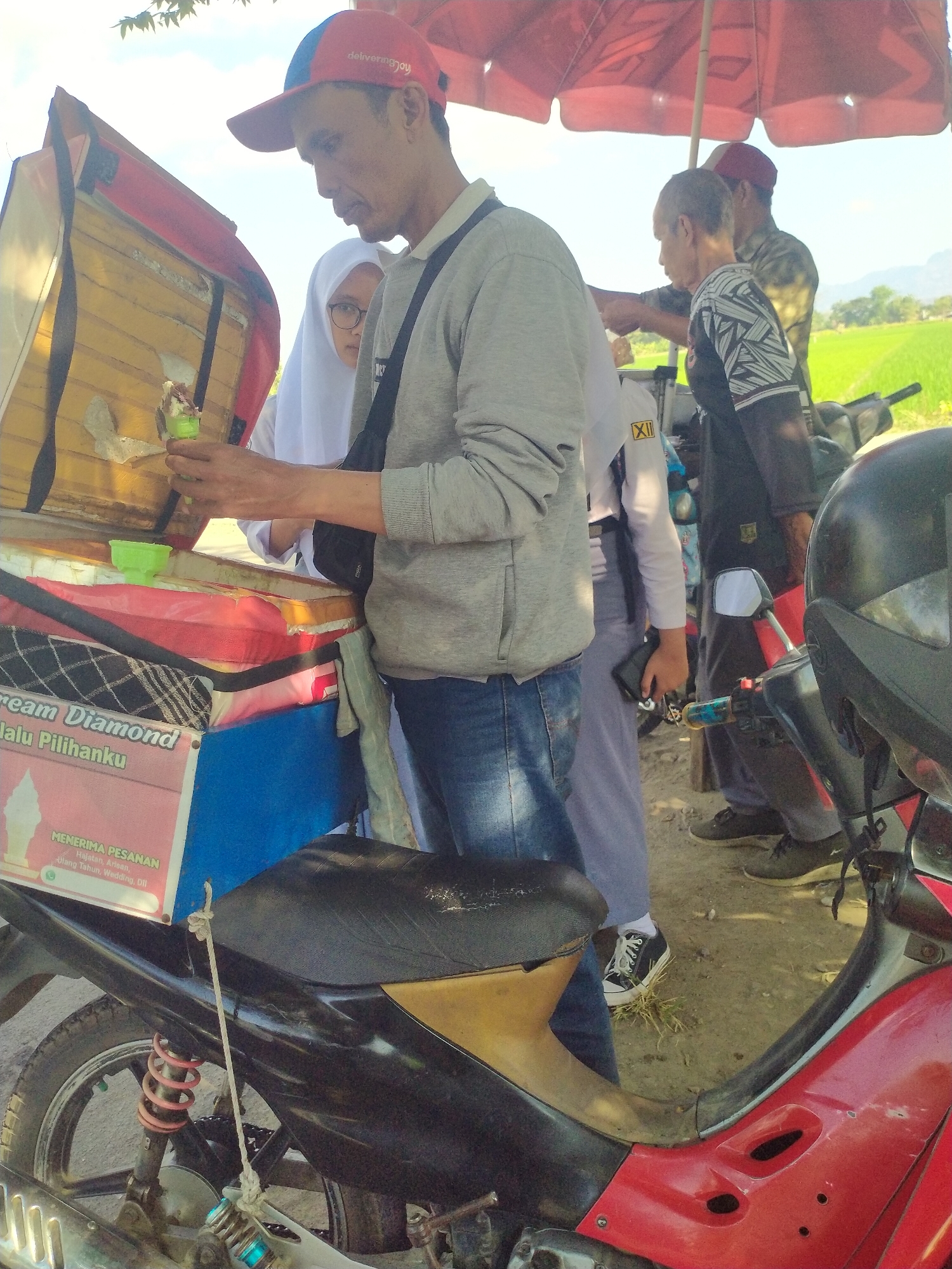 In the city of Begajah, Indonesia, an ice cream vendor wearing a red and blue cap and a grey long-sleeved shirt prepares an ice cream cone. He stands beside a red and yellow ice cream cart attached to a red motorcycle. A young woman in a white hijab and glasses observes from behind the vendor. Behind them, a man in a camouflage shirt and a man in a black shirt with a geometric pattern stand under a red umbrella. The background reveals a bright sky and a lush green rice field, typical of the rural landscape in Begajah. The scene captures a moment of commerce amidst a serene, natural environment.