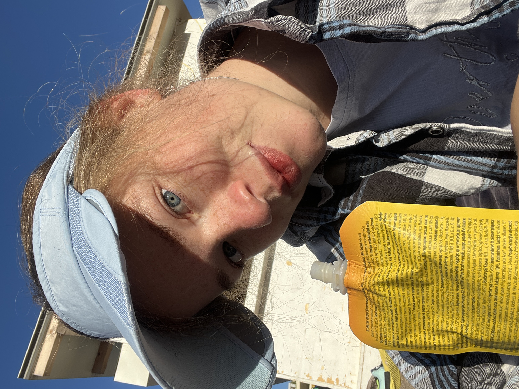 The image shows a close-up, low-angle selfie of a young person with fair skin, blonde hair, and blue eyes, wearing a light blue baseball cap and a plaid shirt over a light-colored t-shirt. The person is holding a yellow pouch with text printed on it. The background shows a bright blue sky and a corner of a white building. The text on the pouch is in a language other than English. No specific location in Bečići, Montenegro, is discernible from the image alone.