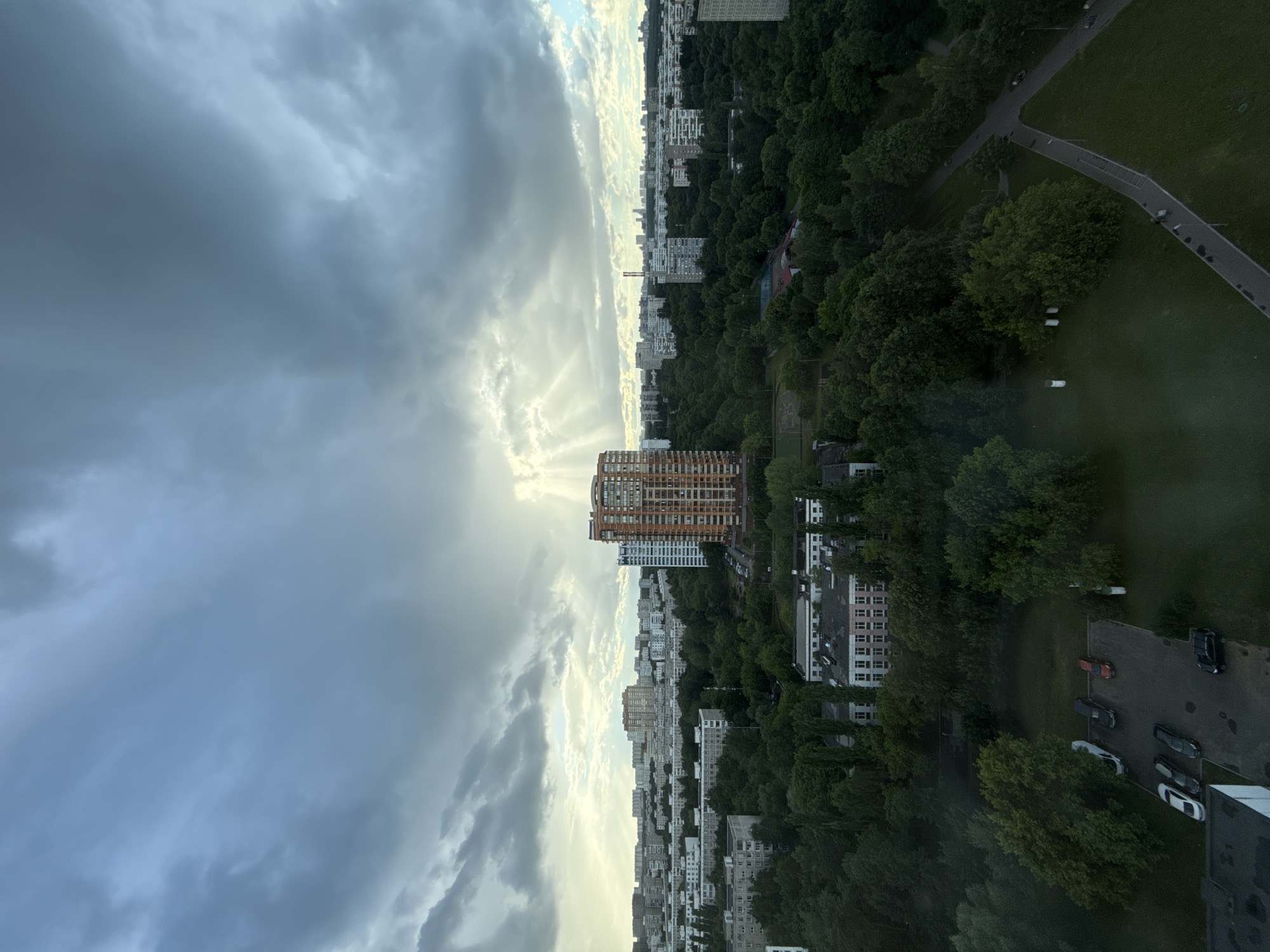 This is an aerial, wide-angle photograph looking out over a densely populated residential area in Moscow, Russia. The sky is dramatic and overcast with bright rays of sunlight piercing through the clouds in the center of the frame, creating a crepuscular effect. In the foreground, a patch of grass and what appears to be a rooftop of a building are visible.
Dominating the mid-ground is a cluster of apartment buildings and lush, dense green trees that form a significant portion of the landscape. A distinct, tall, orange-colored apartment building with many windows stands out in the middle of this green expanse. Several other multi-story residential buildings, varying in color and architectural style, are scattered throughout the scene, interspersed with the trees. A parking lot with several cars is visible near the base of the orange building. A paved pathway winds through the greenery, with a few figures walking on it. In the background, the cityscape stretches out, a sea of grey and white buildings under the brooding sky. The overall impression is one of a large urban environment, possibly a park district, with a mix of residential structures and abundant natural foliage.