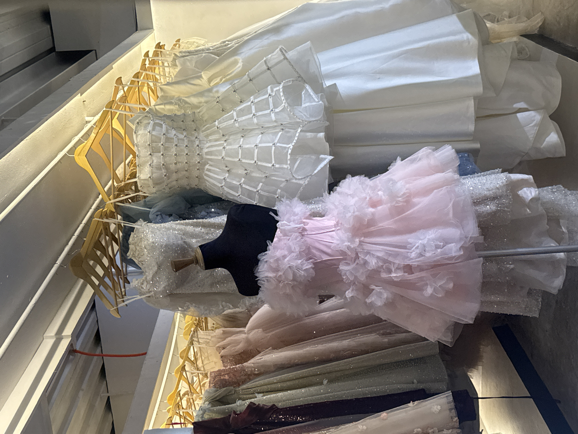 The image depicts a retail display of formal dresses, likely in a boutique or a dress shop. Several dresses are hung on wooden hangers from metal racks, and one dress is displayed on a black mannequin. The dresses vary in color and style, with noticeable examples including a voluminous light pink dress adorned with floral appliques, and a white strapless dress with a structured bodice and a tiered, cage-like skirt. Other dresses visible are in shades of white, cream, green, and possibly burgundy, with some appearing to be embellished with sequins. The background shows a ceiling with exposed metal beams and lighting fixtures, suggesting an indoor commercial space. The overall impression is that of a curated collection of evening wear or bridal gowns. There is no discernible evidence to place this scene specifically in Davao City, Philippines, as the visual cues are generic to a dress shop environment.
