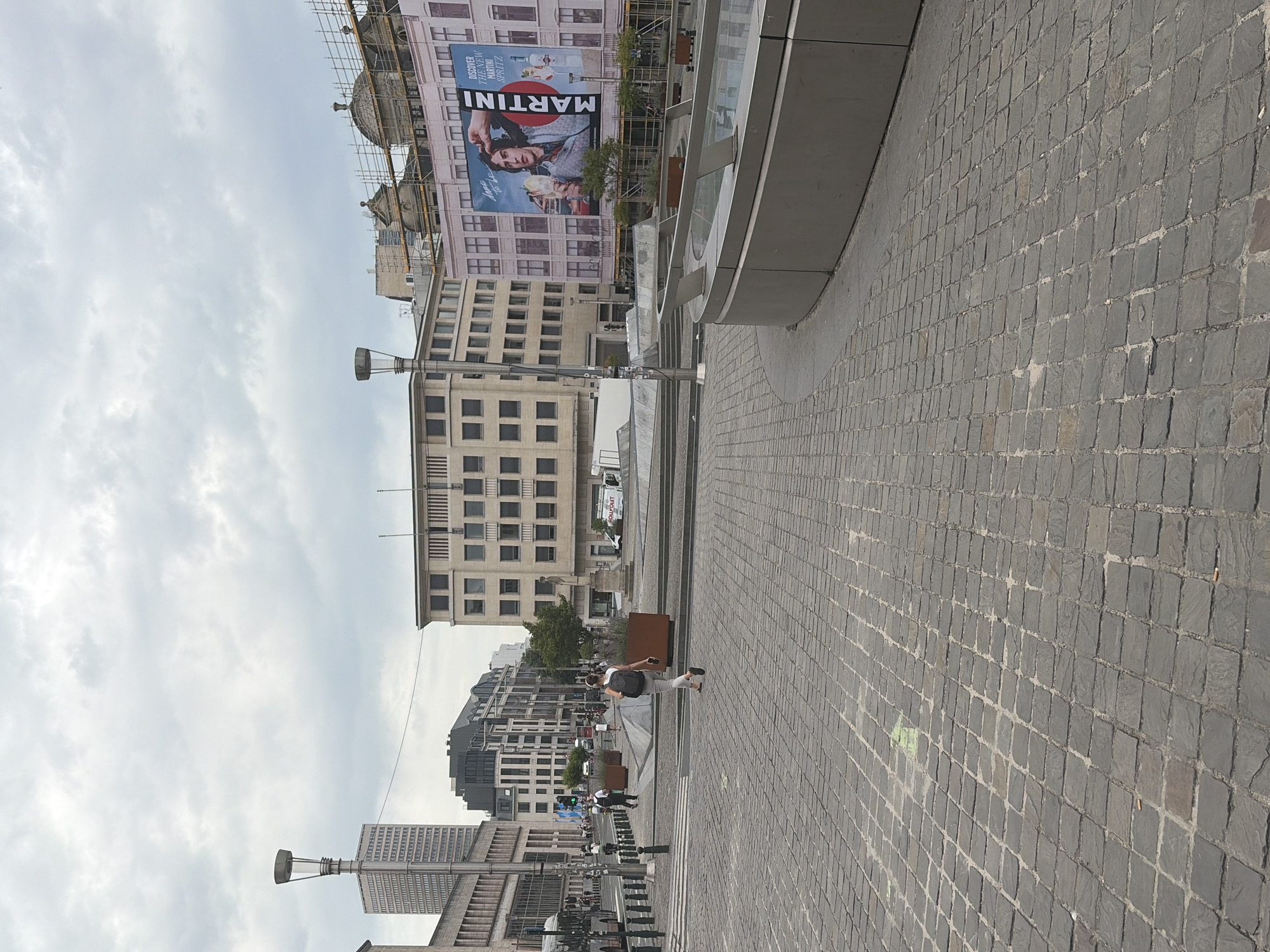 In a public square paved with cobblestones in Brussels, Belgium, a person wearing a backpack walks away from the camera. They are wearing a white t-shirt and light-colored pants. To their left, a street with bollards and a bus is visible, with a tall, modern building in the background. To their right, a large billboard advertising Martini Spritz featuring a man holding a drink is mounted on a building undergoing scaffolding. Other buildings with a mix of architectural styles are also present. A circular fountain structure is in the foreground to the right, and trees are interspersed throughout the square. The sky is overcast with gray clouds.