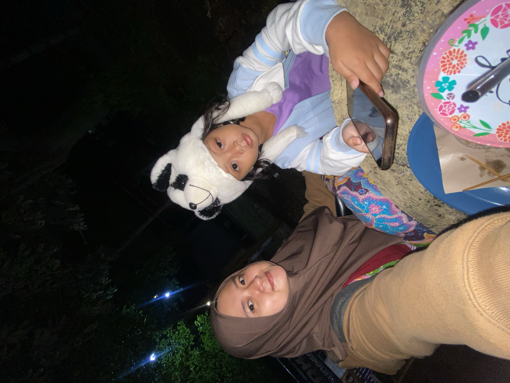 Two females are shown in an outdoor setting in Kalitengah, Indonesia. The younger female is wearing a panda hat, a light purple shirt, and a white and blue hooded sweatshirt. She is holding a brown cellphone. The older female is wearing a brown hijab and a tan-colored jacket. There is a circular pink floral patterned serving tray and a blue plate with food and disposable wooden skewers. Vegetation and a streetlight are visible in the background.
