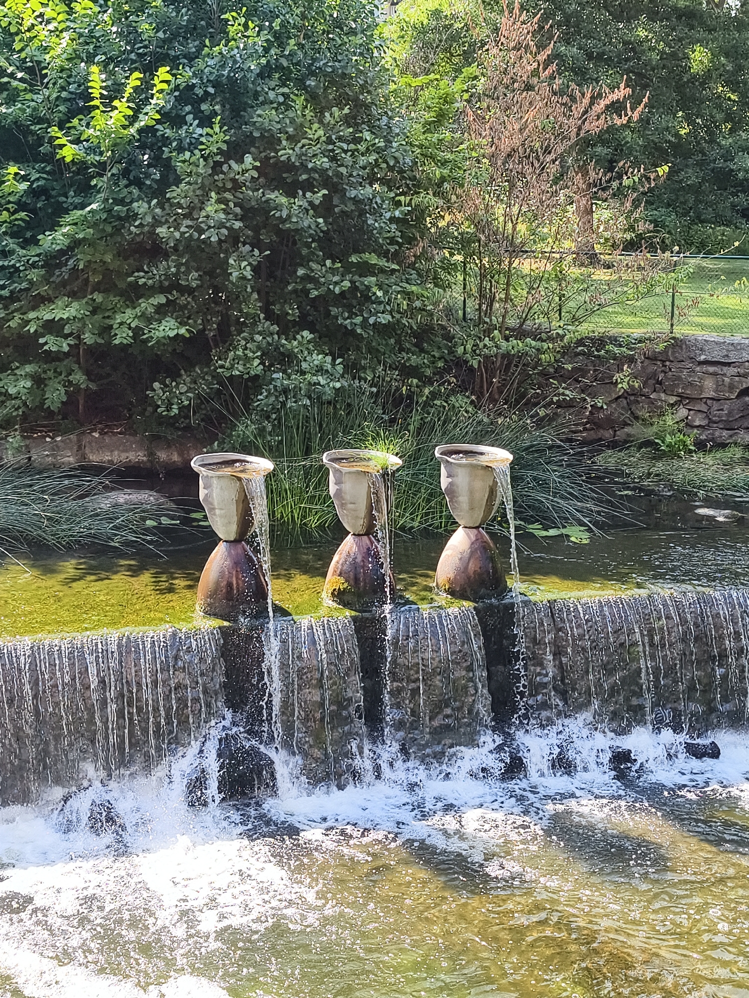 The image depicts a three-tiered water fountain feature in a park-like setting. Each tier consists of a dark, conical base topped with a light-colored, bulbous vase. Water flows from the top vase of each tier, cascading down to the next, and eventually over a rough-hewn stone weir into a body of water below. The weir itself creates a small waterfall with frothy white water.
Surrounding the fountain are lush green trees and vegetation. In the background, a stone wall runs horizontally, with more greenery behind it. The water in the foreground has a yellowish-green tint and reflects the sunlight. There are no people or animals visible in the image. The overall scene suggests a tranquil outdoor space. This water feature is located in the Valla recreation area in Linköping, Sweden.