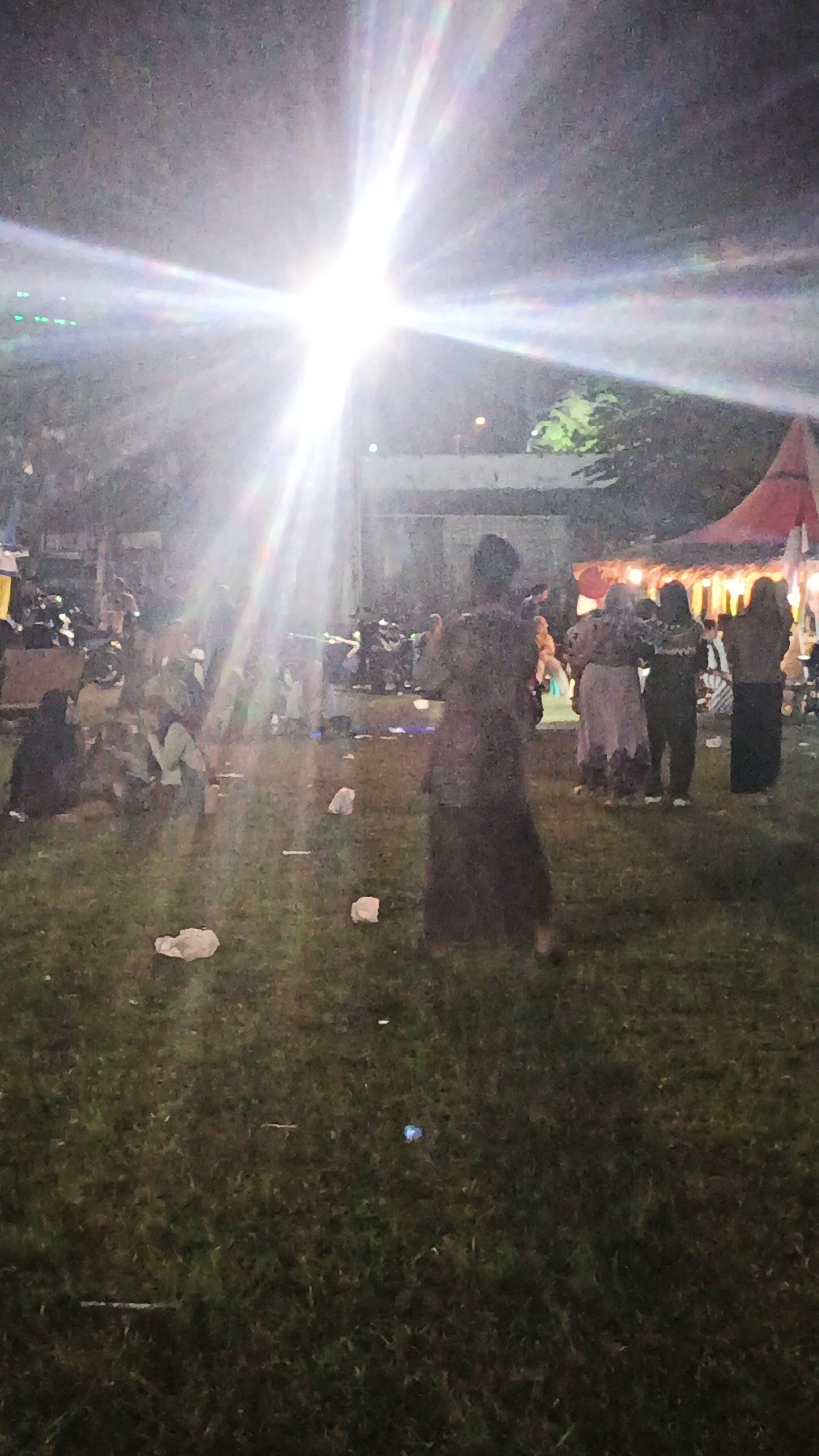 The image depicts an outdoor scene at night, possibly a gathering or event, in Ketemasdungus, Indonesia. A bright, lens-flared light source dominates the upper center of the frame, casting strong beams and creating a starburst effect. In the foreground, a grassy field is visible with scattered white debris, likely paper or plastic. Several people are present throughout the image. In the mid-ground, a group of individuals are seated or standing on the grass, some appearing to interact. A figure in a dark, flowing dress stands prominently in the right mid-ground, facing away from the camera. In the background, more people are visible, some near parked motorcycles or scooters. To the far right, a red tent or canopy is partially visible, illuminated from within, suggesting a structure associated with an event. The overall impression is of a lively, outdoor nighttime gathering.
