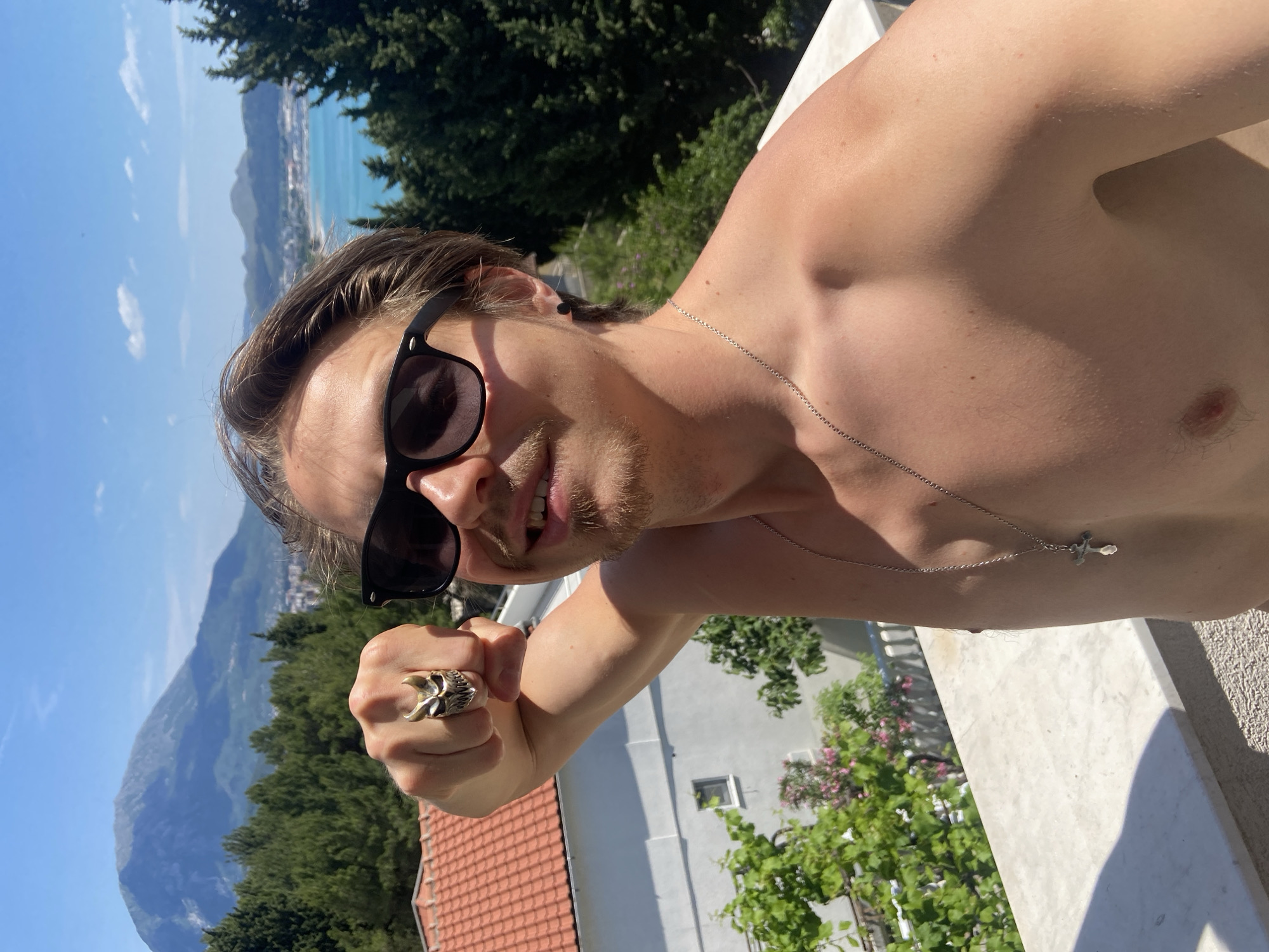 A fair-skinned man with brown hair and sunglasses is taking a selfie outdoors in Šušanj, Montenegro. He is shirtless and wearing a silver necklace with a cross and a skull ring on his raised fist. In the background, a landscape with a mix of trees, rooftops, mountains, and a body of water is visible under a clear blue sky. The presence of a stone railing suggests he is on a balcony or elevated platform.