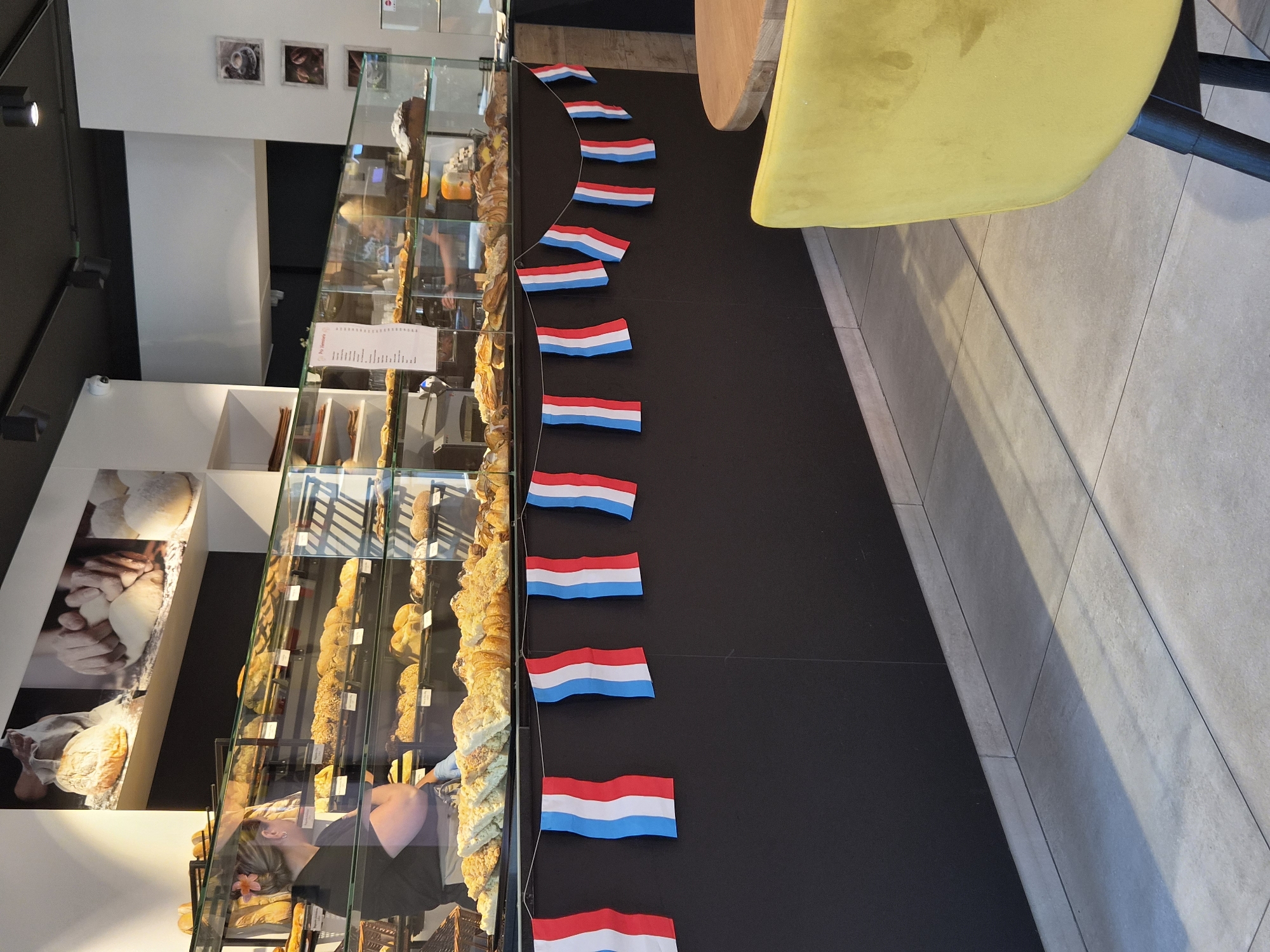 A medium shot captures the interior of a bakery, with a focus on the display of baked goods and a decorative string of French flags. In the background, a bakery employee with blonde hair pulled back and a pink flower accessory is visible, wearing a black t-shirt and a white apron. They are standing behind a glass display case filled with various pastries, croissants, and bread. The display case is well-lit, showcasing the products on multiple shelves. Above the display, a large black and white photographic mural depicts hands kneading dough and loaves of bread. A string of small French flags, red, white, and blue, is draped across the front of the counter, adding a festive touch. The lower portion of the image shows a black wall and a light-colored tiled floor. In the foreground, to the right, a corner of a bright yellow upholstered chair is visible, and a portion of a round wooden table. The overall scene suggests a commercial establishment, likely a bakery in Sanem, Luxembourg, given the presence of French flags.