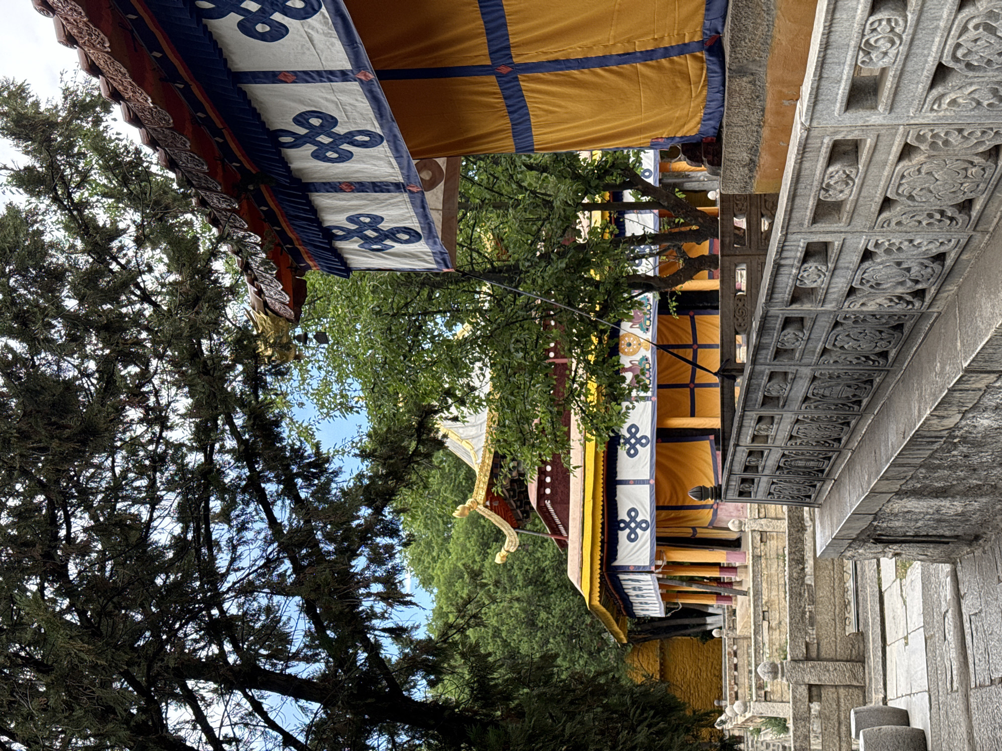 The image shows an upward perspective of a building in Chengguan District, China. The architecture is decorated with Tibetan motifs, specifically with yellow and white awnings with blue and red detailing. The stone railing is decorated with carved floral patterns. Green trees are interspersed along the building.