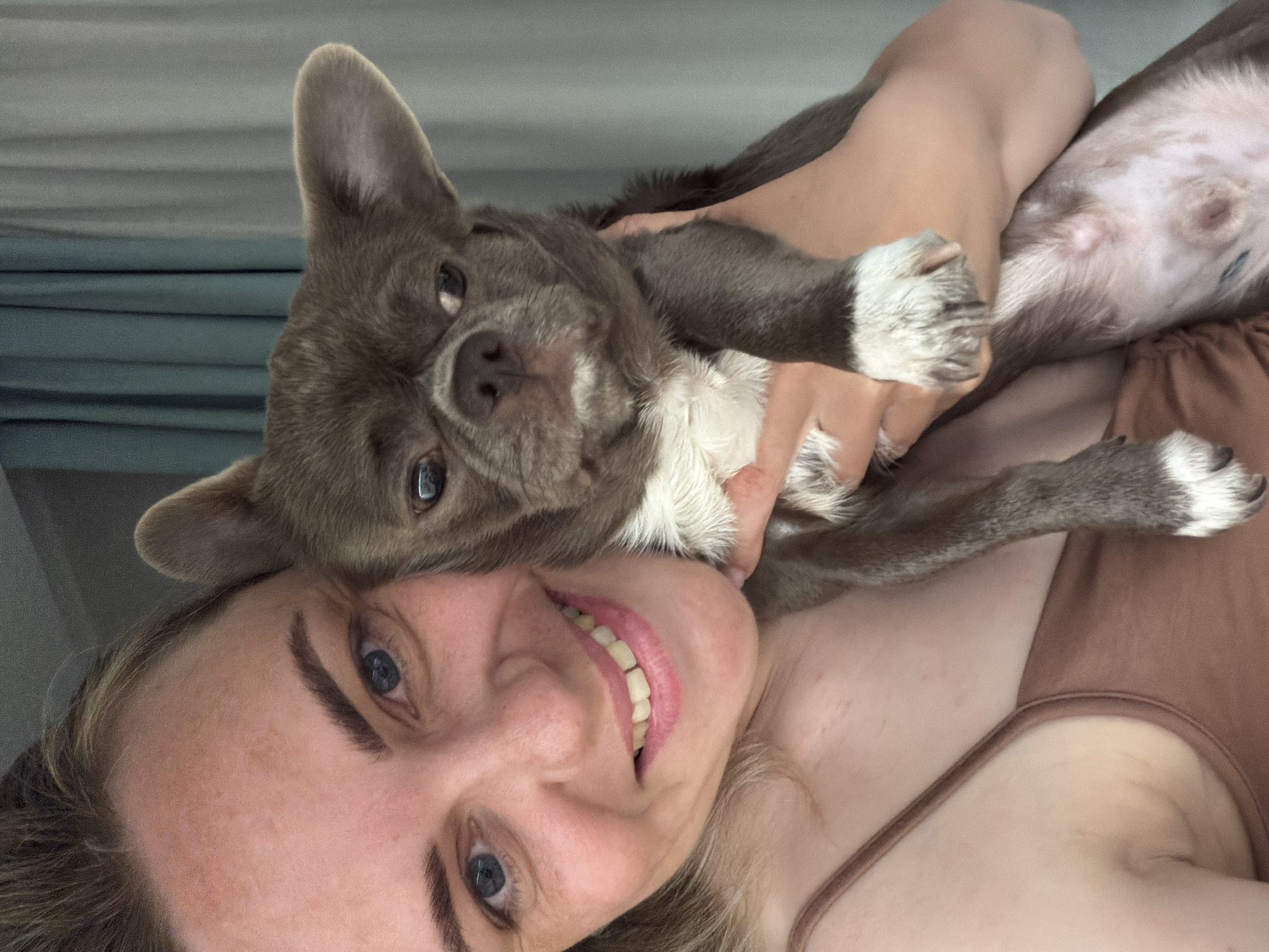 A woman with blue eyes and fair skin is smiling directly at the camera. She has a brown tank top on. A gray dog is lying on her chest, being held by her arms. The dog has white paws. The background appears to be a gray wall. The image does not appear to be taken in Votkinsk, Russia, as there are no indicators of location.