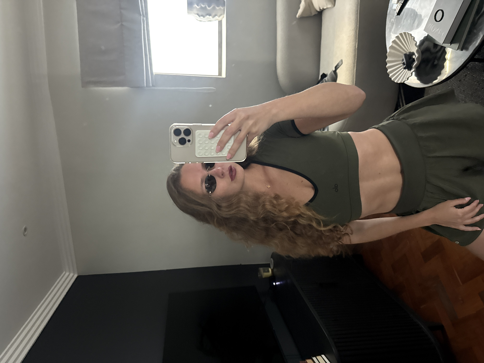 A woman is taking a mirror selfie indoors. She is wearing sunglasses and an olive-green workout set with a bra top and shorts. The setting includes a window with grey curtains, a grey sofa, and a black and silver coffee table. There is also a black wall or TV screen partially visible in the lower left corner. The background elements suggest a modern interior. The specific location in Cape Town, South Africa is not discernible from the image.