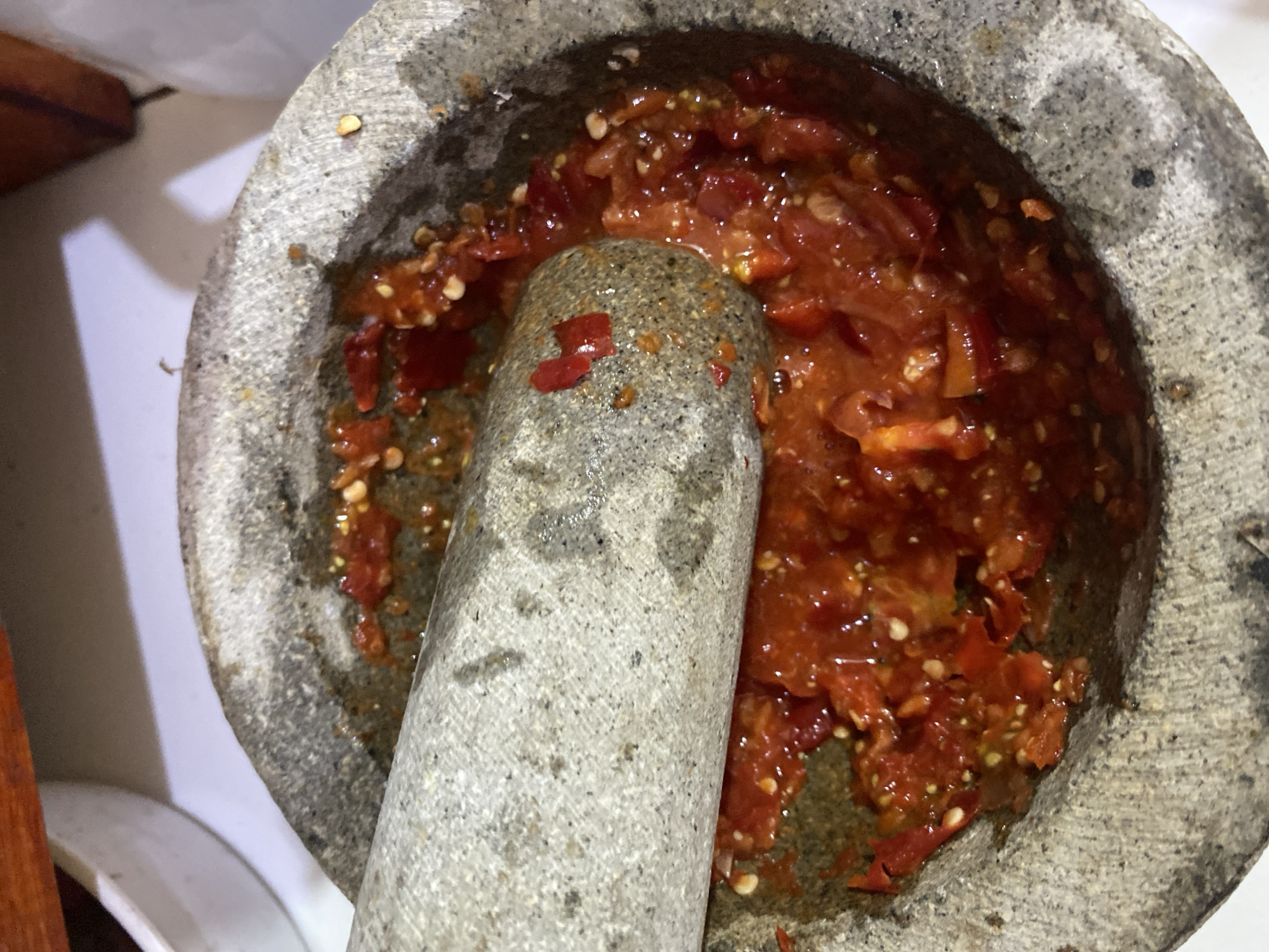 A top-down, close-up view shows a stone mortar and pestle with a vibrant red, chopped chili paste inside. The mortar is rough and textured, with flecks of lighter and darker gray stone. The pestle, also gray and cylindrical with a rounded end, rests inside the mortar, partially submerged in the chili mixture. The chili paste is wet and appears to be finely chopped, with visible seeds and small pieces of red chili. The background is blurred, with hints of white surfaces and a dark wooden object at the top edge of the frame. The lighting suggests an indoor setting. The image does not contain enough information to determine the specific location in the city of Ta', Indonesia.