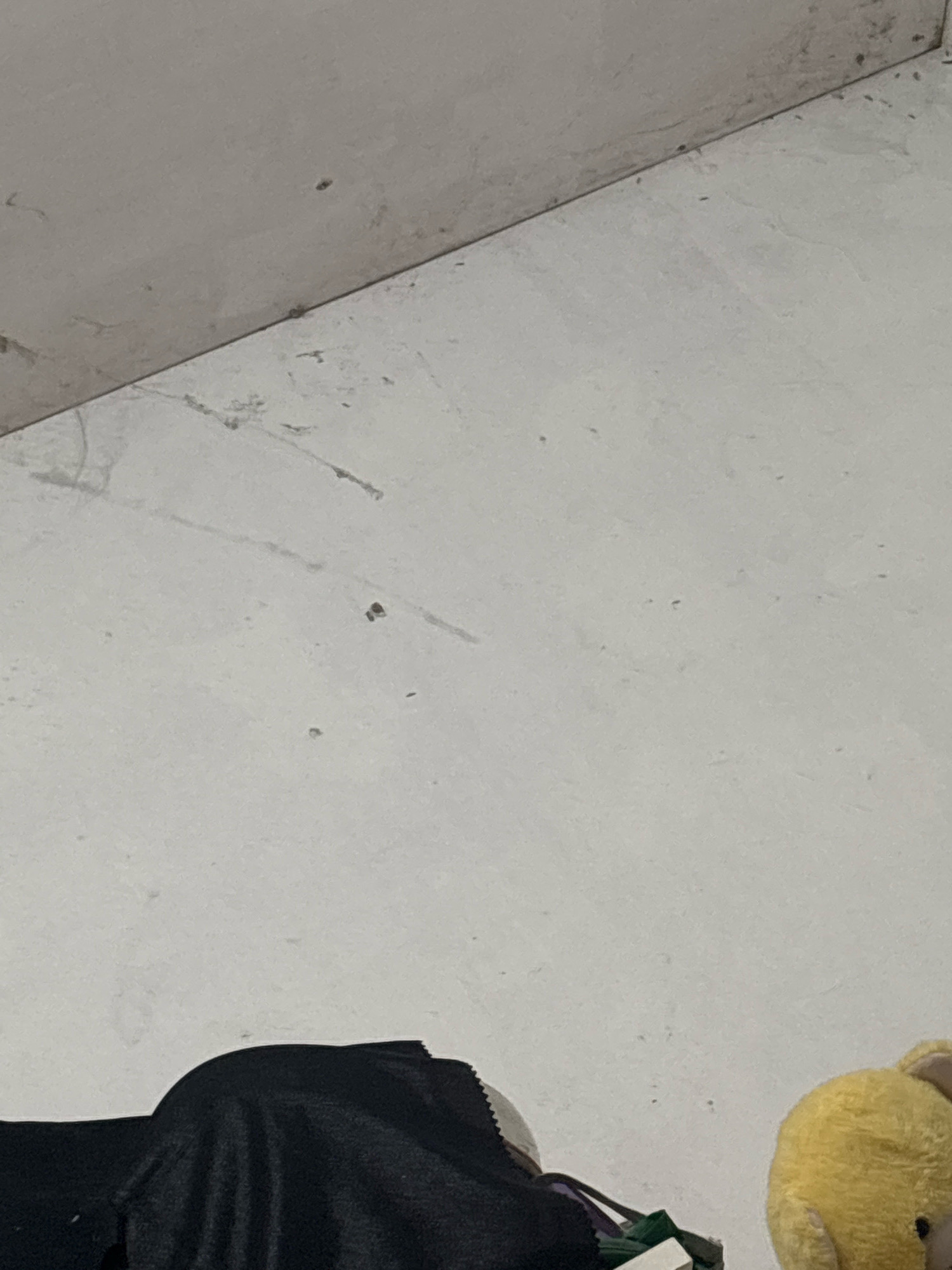 The image depicts an interior scene with a light-colored wall that appears to be plaster or painted concrete. The wall has some scuff marks and dust, suggesting it might be an unfinished or older surface. In the lower portion of the image, a black fabric object is visible, possibly clothing or a bag, with some green and purple straps or material attached. To the right of the black fabric, a portion of a yellow stuffed toy, likely a bear, is visible. The background wall is plain and light-colored, with some faint lines that might be shadows or imperfections in the surface. The lighting appears to be artificial and somewhat dim. There are no people or animals visible other than the stuffed toy. The image quality is moderate, with some noise and a slight blur. The location is described as North Jakarta, Indonesia, but the image itself does not contain any specific visual cues to confirm this geographical context.