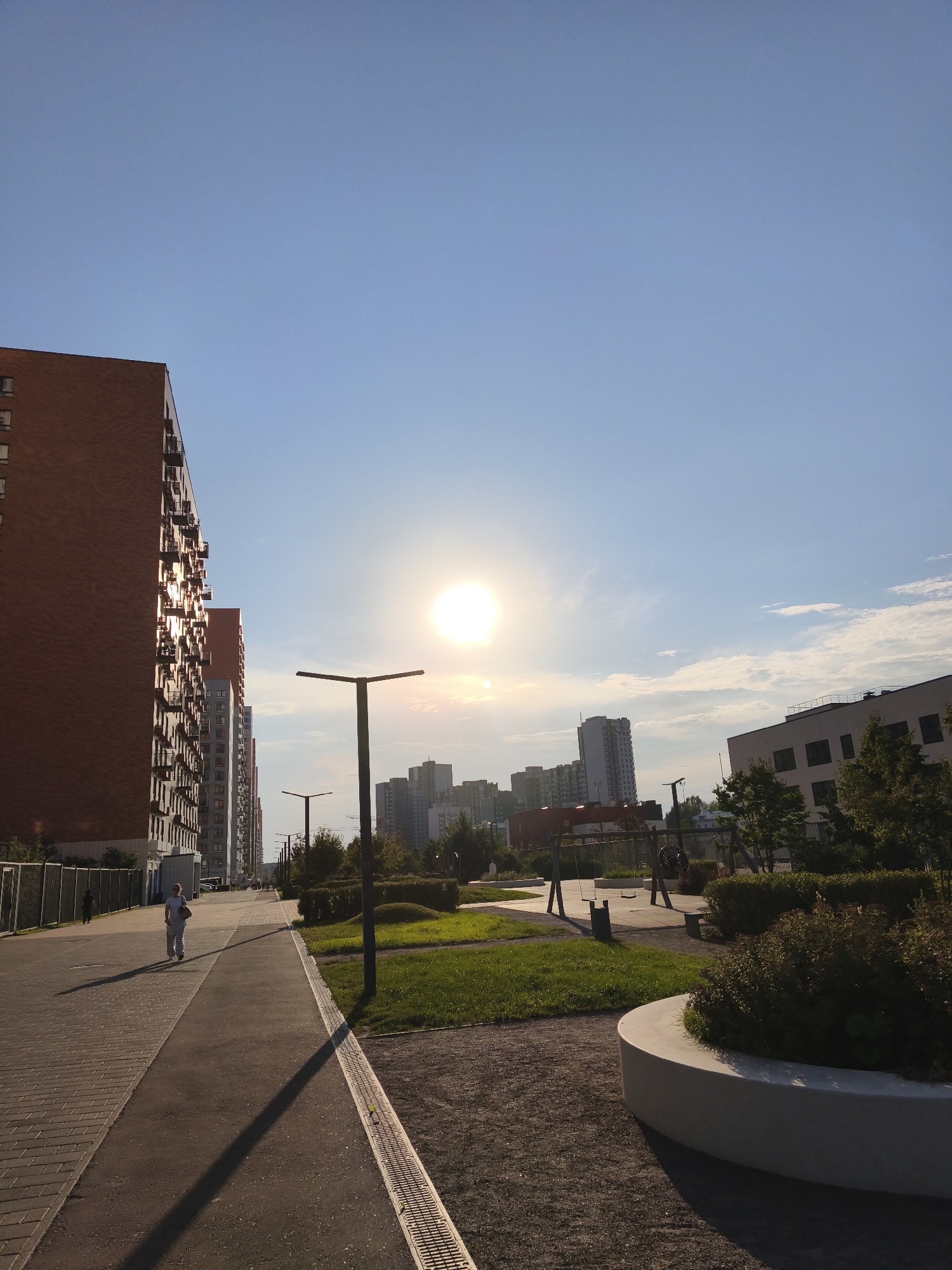 An outdoor scene captures a modern urban environment under a clear blue sky with the sun prominently visible. The perspective is from a pedestrian walkway, leading towards a backdrop of several residential buildings and a playground.
In the foreground, a man is walking away from the camera on a paved sidewalk. The sidewalk is divided into two sections: one paved with rectangular bricks and the other with asphalt. A long shadow is cast by the man and a streetlamp onto the walkway. To the right of the sidewalk, a patch of well-maintained green grass is visible, bordered by a low, curved concrete planter filled with bushes. A modern playground with swings is situated further back, with some children visible on the equipment.
The mid-ground features a row of tall, brick-faced apartment buildings with numerous balconies. Beyond these, a cluster of taller, more modern skyscrapers form the distant city skyline. Streetlights with a minimalist design are placed along the walkway.
The overall impression is that of a sunny day in a newly developed residential area, likely within Moscow, Russia, given the architectural style of the buildings and the general urban planning visible. The environment appears clean and orderly, suggesting a contemporary residential complex.