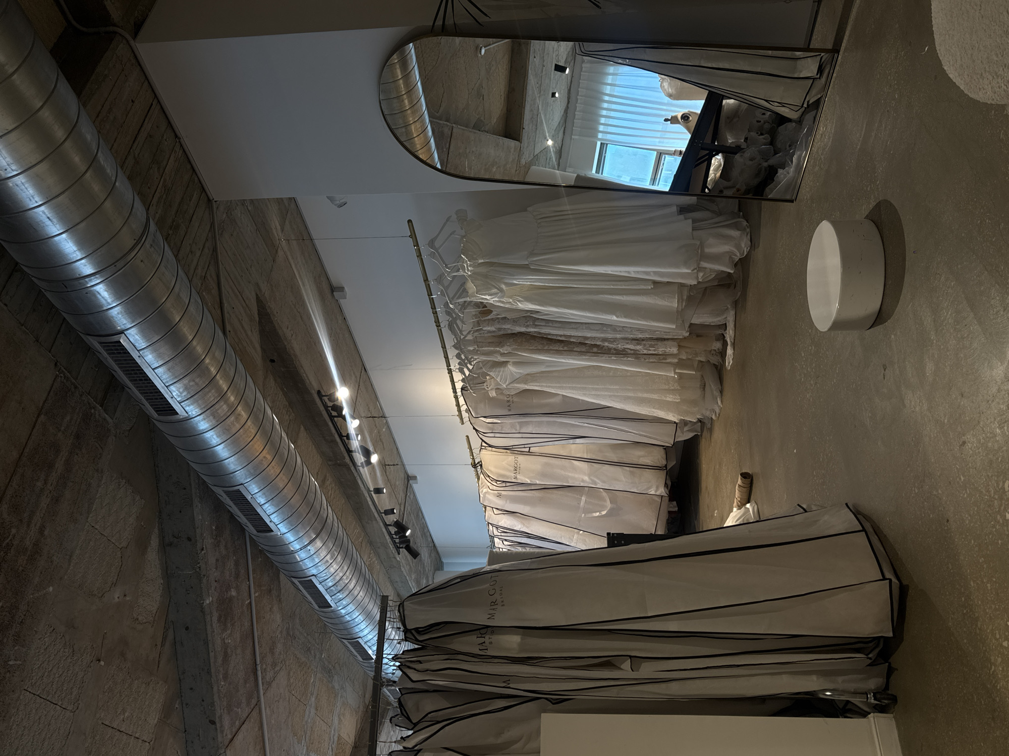 The image depicts the interior of what appears to be a bridal shop, specifically a section with wedding dresses. The scene has an industrial aesthetic, evidenced by the exposed concrete walls and ceiling, along with a large, silver cylindrical duct running horizontally along the ceiling. Below the duct, a rack of wedding dresses covered in white garment bags hangs from a rod. Some dresses are partially visible without the bags, appearing predominantly white with layers of fabric. On the floor, there are stacks of garment bags, likely containing more dresses. A cylindrical white stool sits near the dresses, in an area with a concrete floor. A mirror on the right reflects the store’s interior, including dresses and mannequins, as well as natural light coming from a window.
The store name, "Margot Bridal Studio", is printed on one of the garment bags. The location of the image is likely to be in Tel Aviv, Israel, given the information provided and the style of the studio.